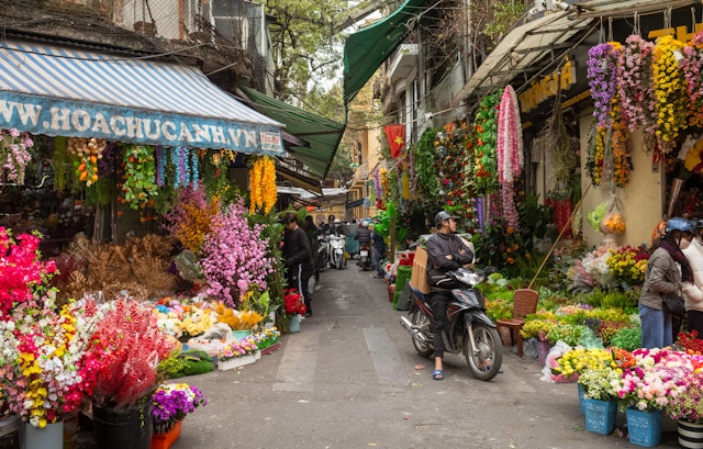 A man on a motorbike waits by shops on either side of an alley selling artificial multi-colored flowers in the Old Quarter of Hanoi, Vietnam