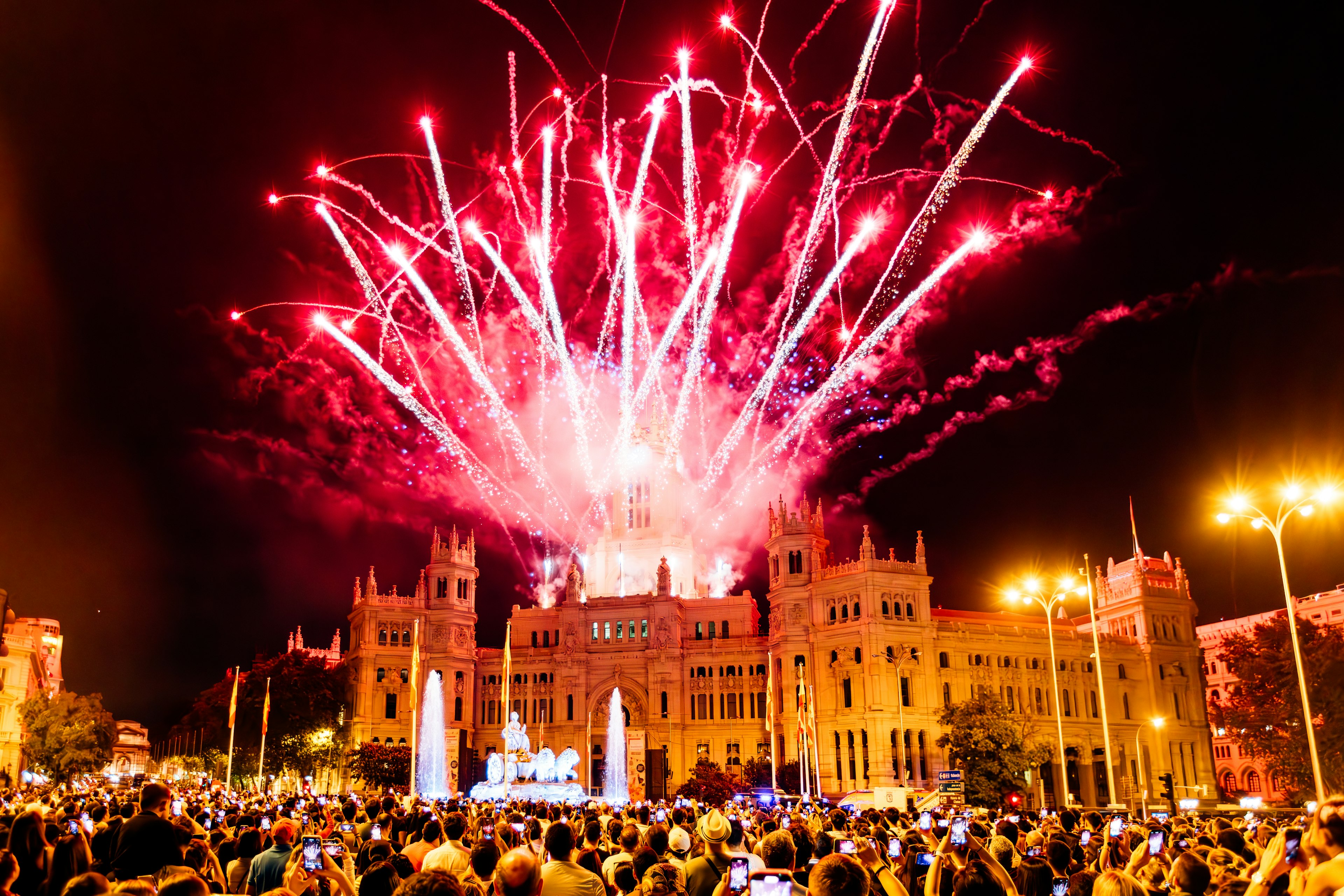 Crowds gather to watch a fireworks display in front of a city building.