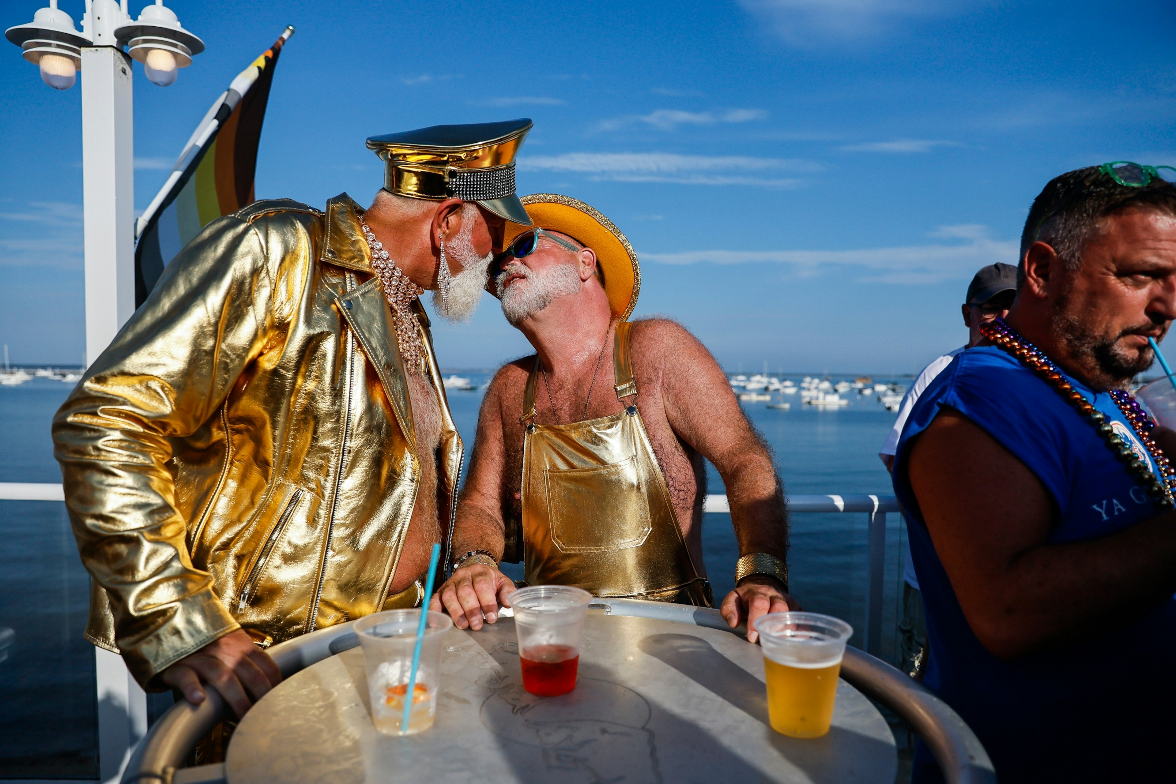 Two men dressed in gold-lamé outfits and sunglasses kiss on a deck overlooking the harbor