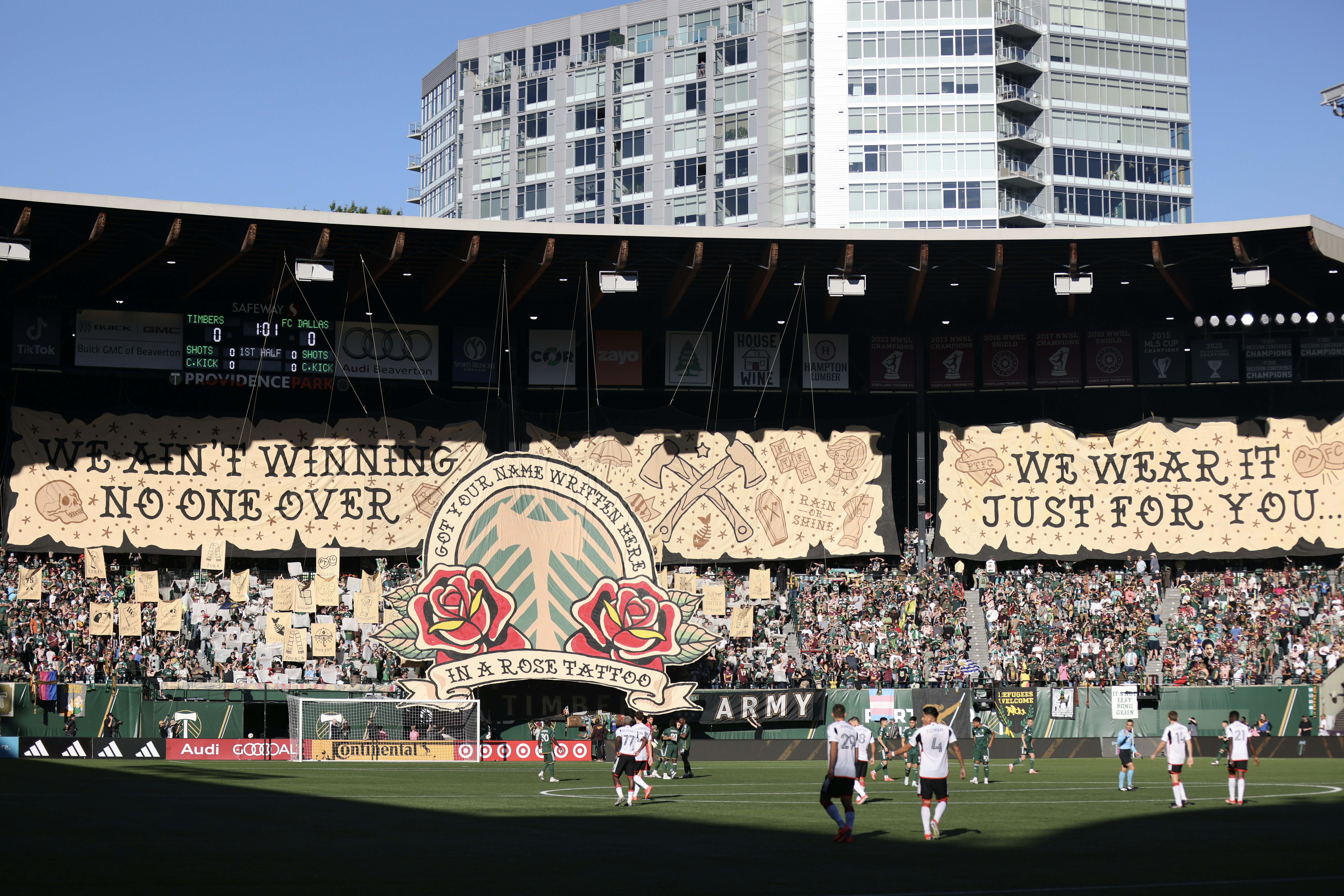 The Timbers Army supporters group displays a tifo before the first half of a game between the Portland Timbers and FC Dallas at Providence Park, Portland, Oregon. Players are on the field, and an apartment tower can be seen behind the stadium.