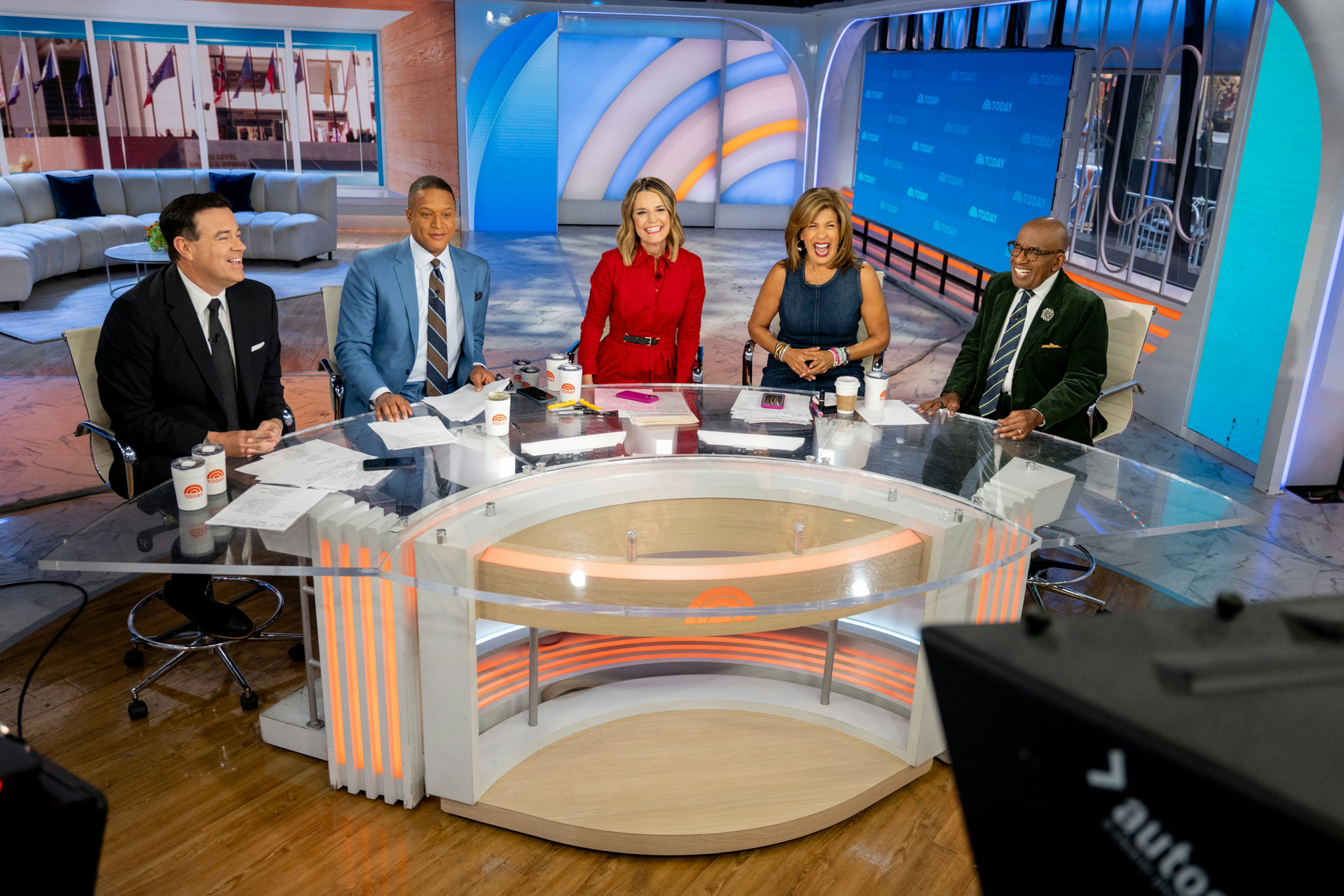 A panel of TV hosts smile as they sit around a large glass desk in a studio