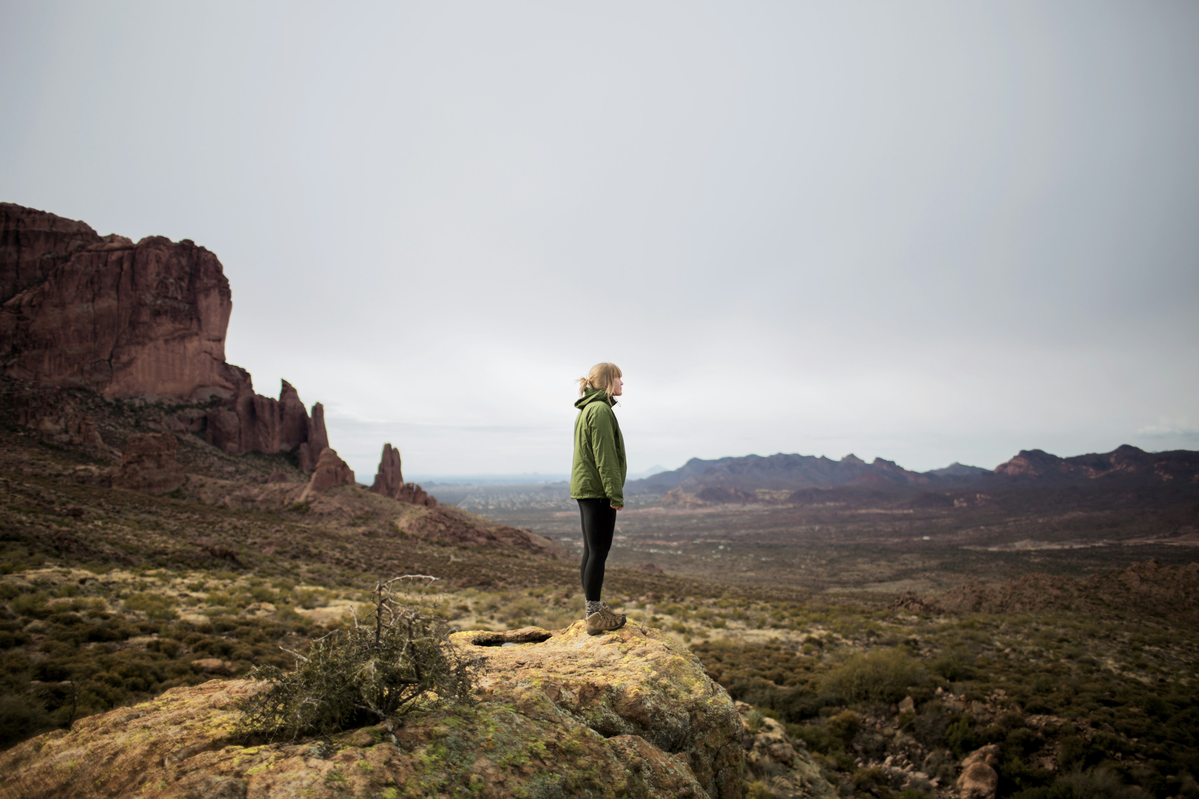 A teenager stands on cliff by rocky mountains against a gray sky, Superstition Mountains, Arizona