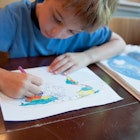 A 6 year old boy is coloring a world map with colored pencils. There is also an atlas on the table. He is concentrating on his geography homework.