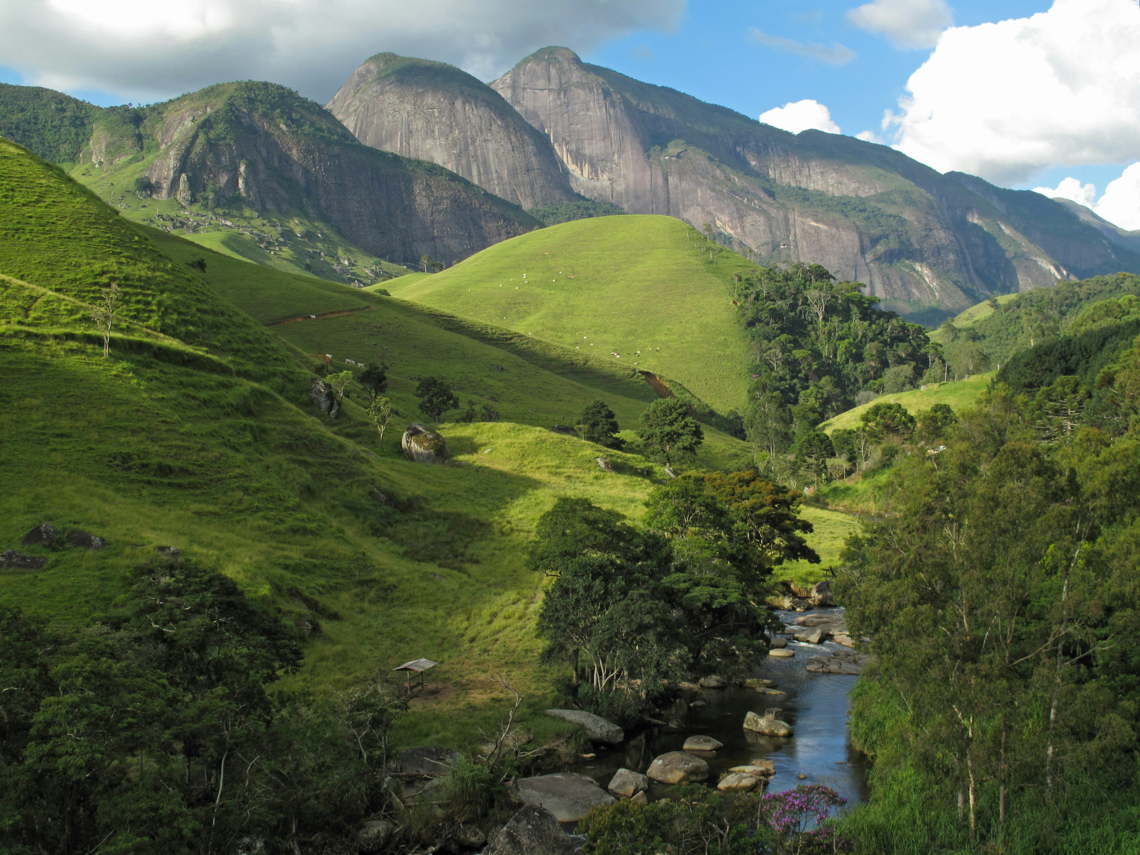 A stream running through rolling green hills and dramatic rocky mountains near the Cachoeira dos Frades waterfall.