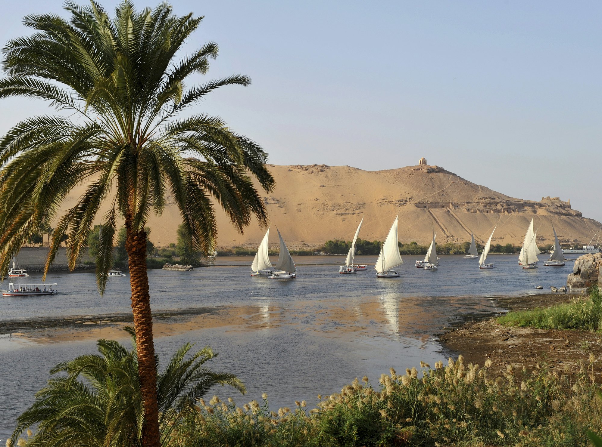 Sailing boats with white sails cruise along a wide river. The tall riverbank opposite has tombs cut into the rock face.