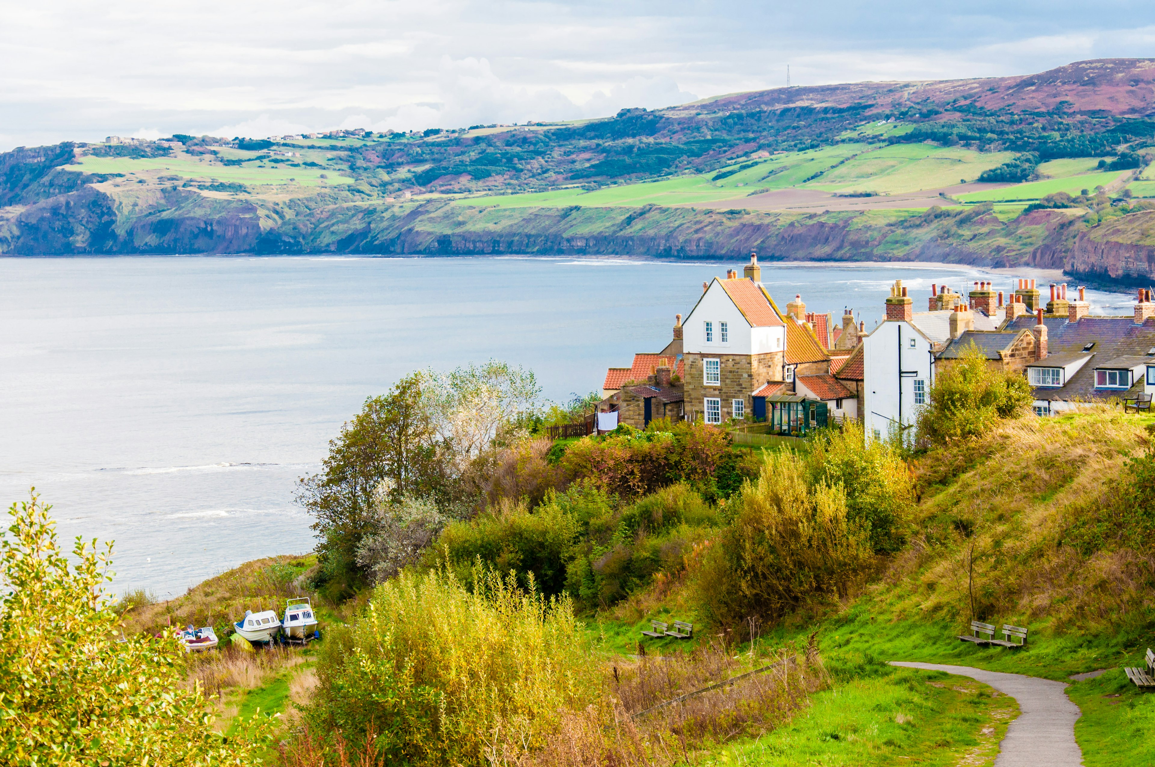 A small path leads to a fishing community with historic white buildings with orange roofs. The town sits on the edge of a bay with rolling green hills in the background. England.