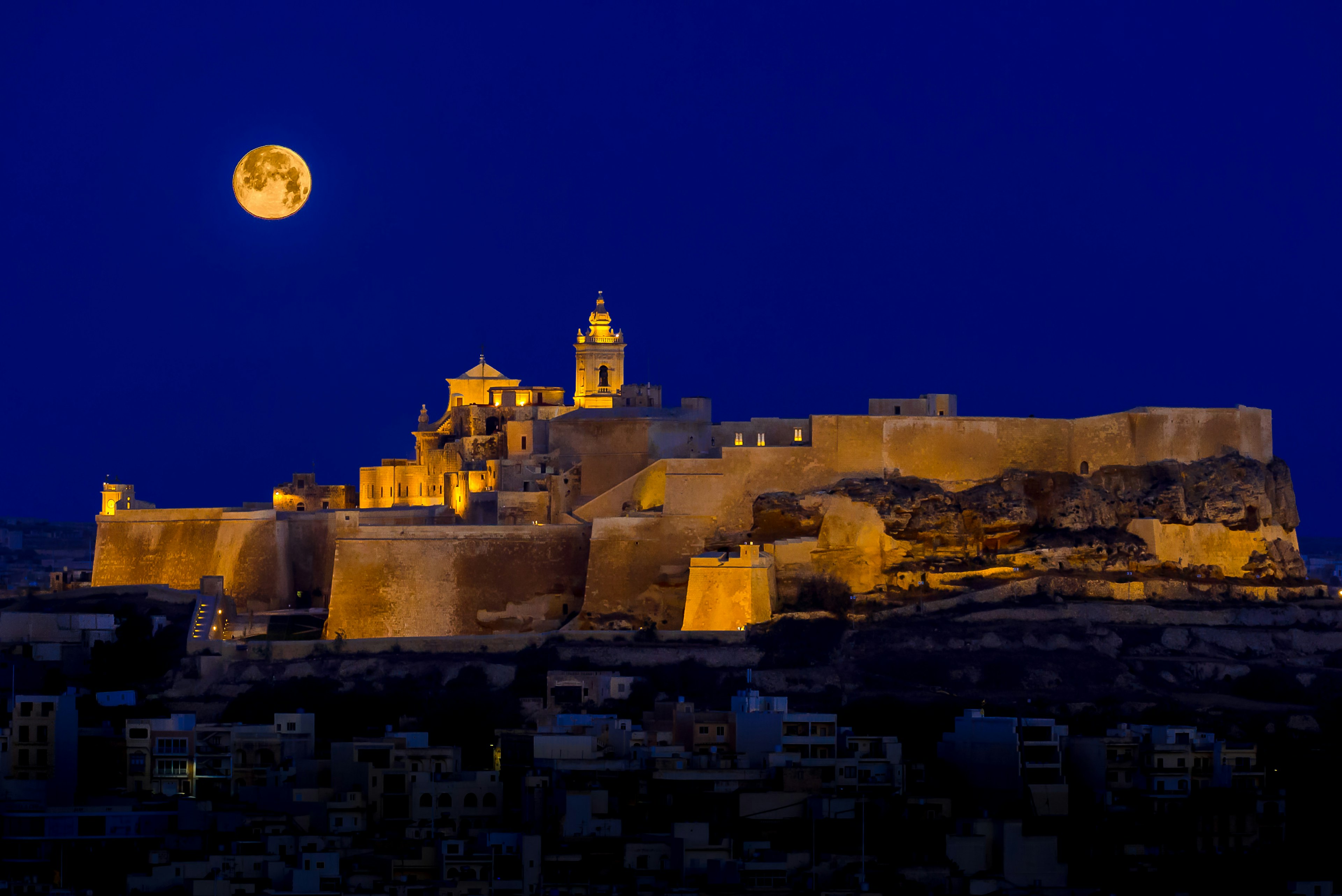 With a full moon rising into a dark sky, the Ċittadella glows gold in floodlights