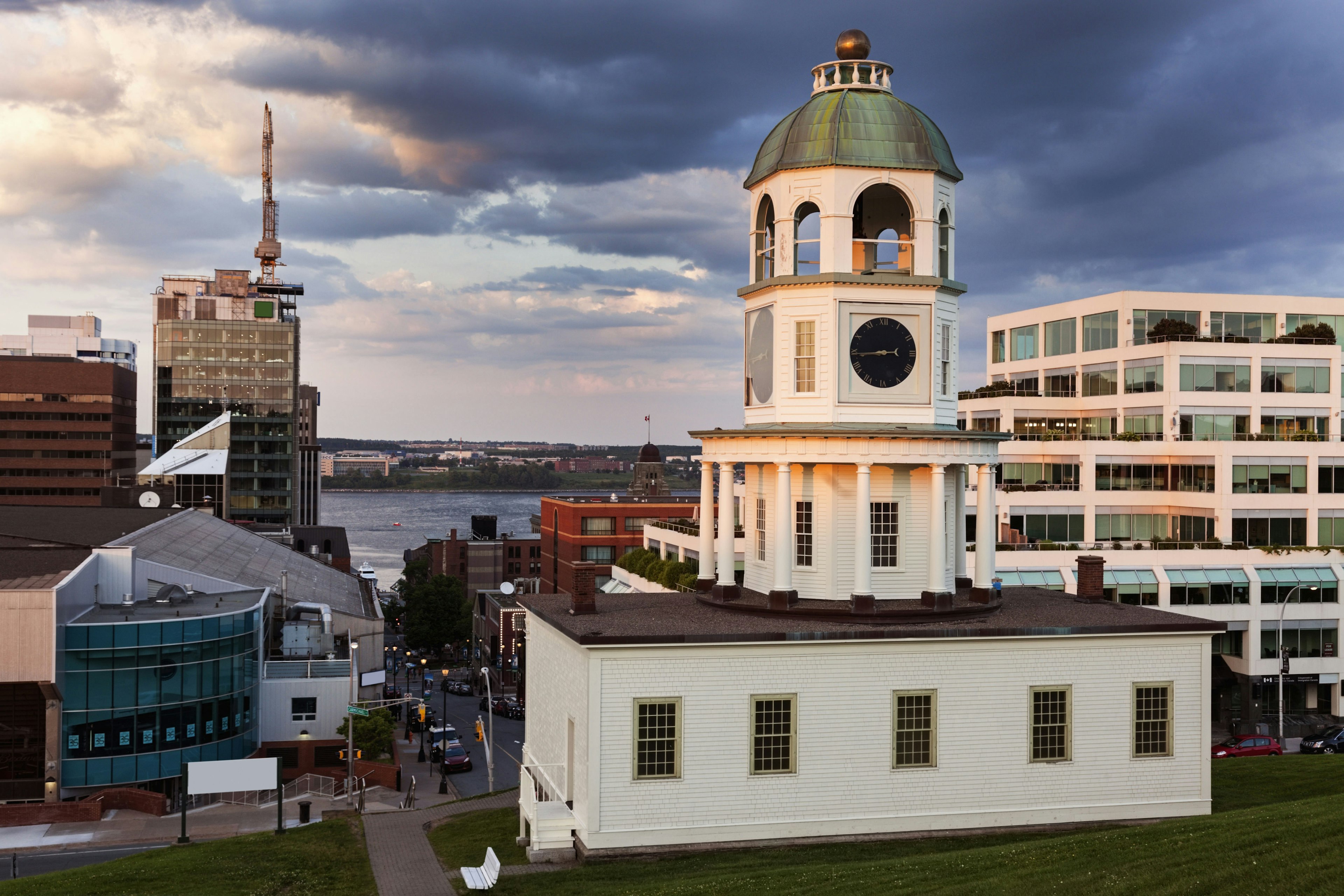 A pinkish glow of sunset illuminates the white siding on a clock tower at the foot of a grassy green hill