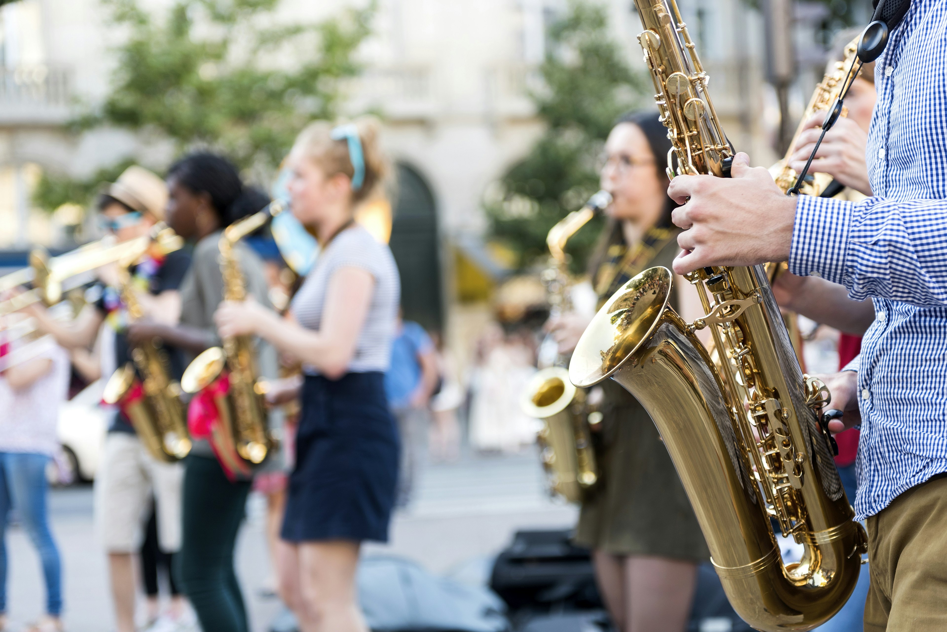 In the foreground is a saxophone and saxophonist, with only the latter's hands and blue-and-white checked shirt torso visible. In the background are brass-toting jazz musicians who are out of focus.