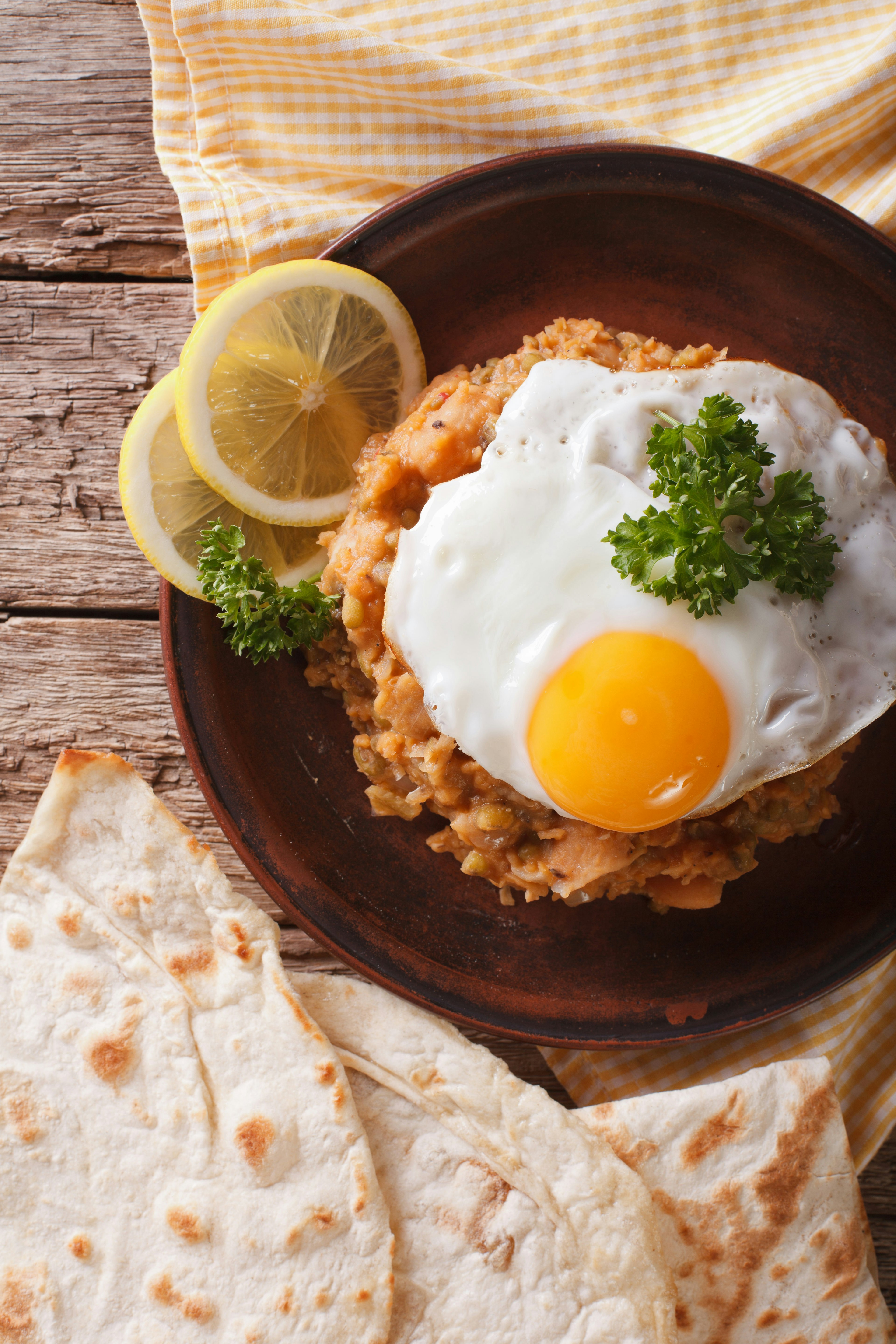 Aerial view of fuul medames with a fried egg and bread on a brown plate garnished with lemon slices