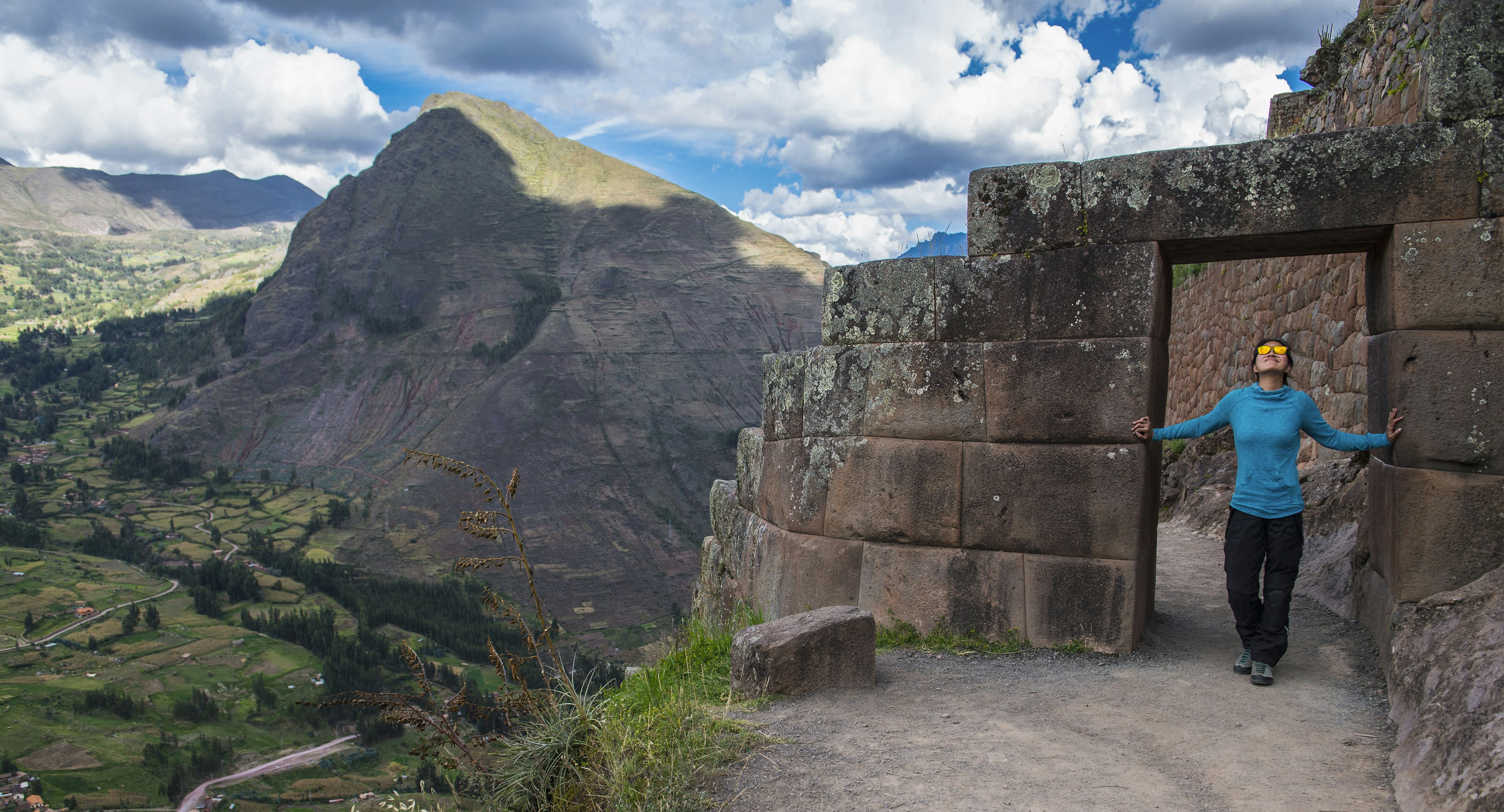 A woman stands in a stone doorway overlooking a green valley next to a rocky mountain