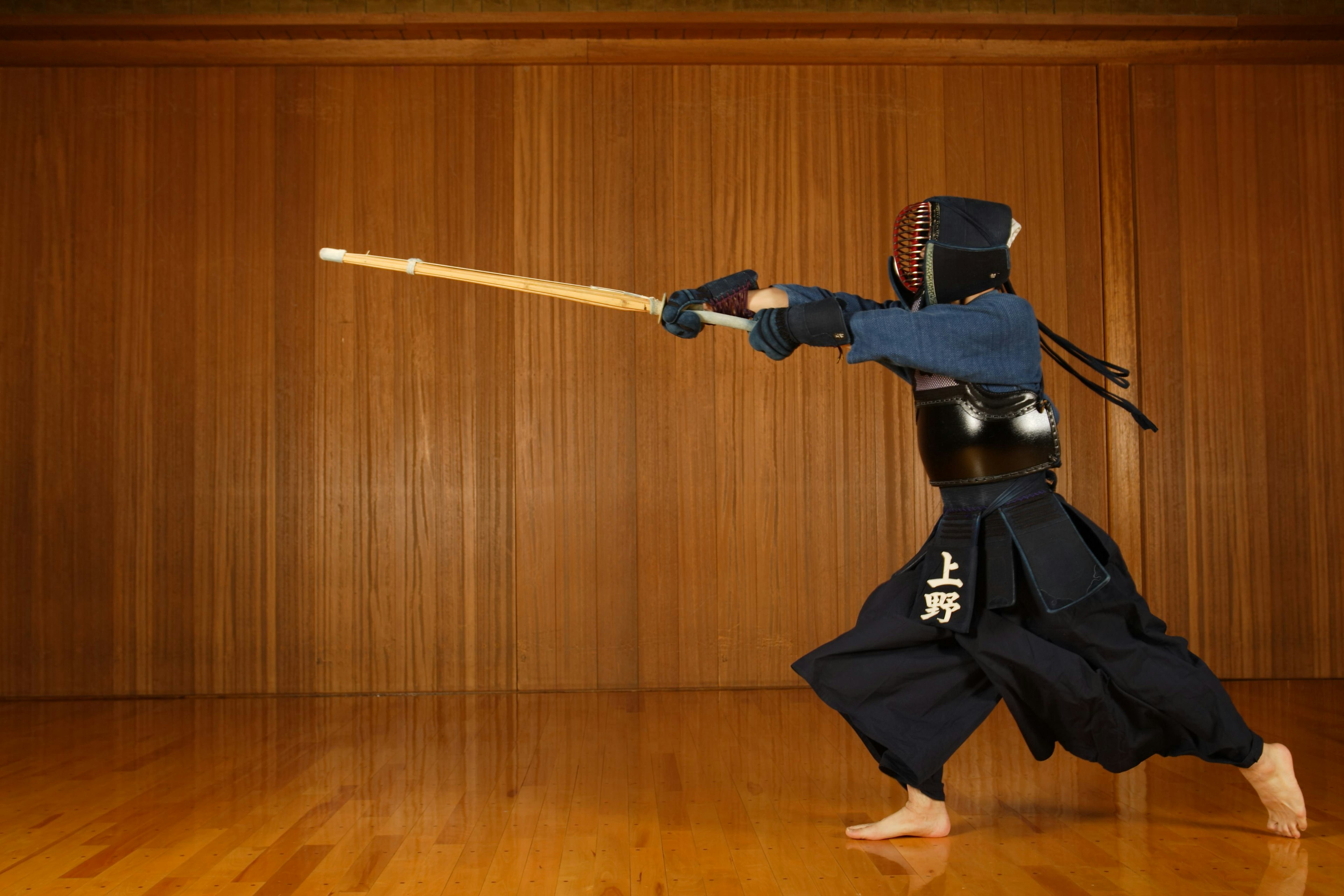 A person dressed in full kendo gear has a fighting stance and holds out the wooden sword