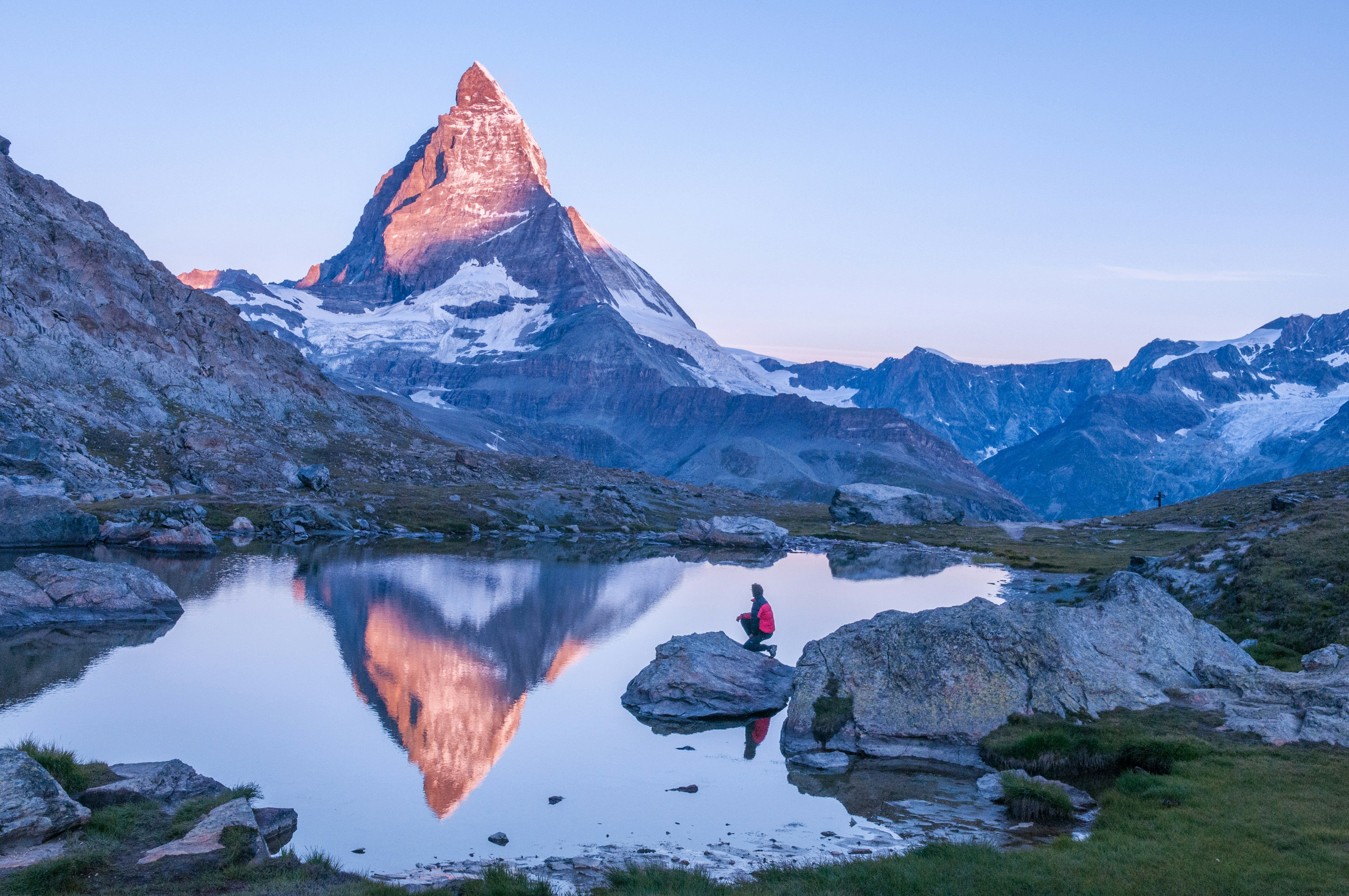 Male hiker kneeling on a rock near a small lake while light from the rising sun strikes the peak of the Matterhorn.