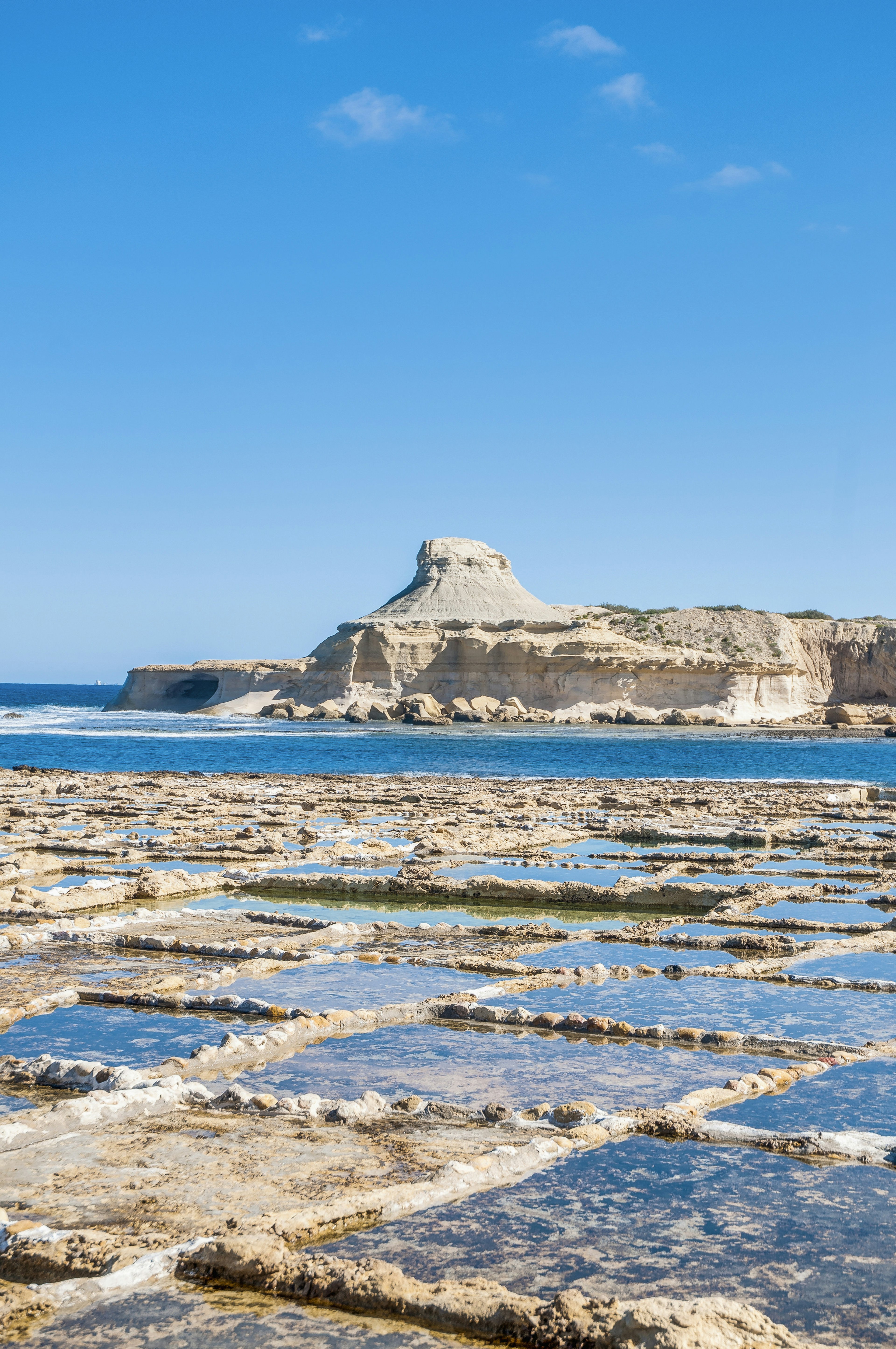 Small, shallow rectangular ponds hewn into the flat limestone bed are filled with water. The sea lies beyond, as does an pyramidal rock outcrop of bleached stone