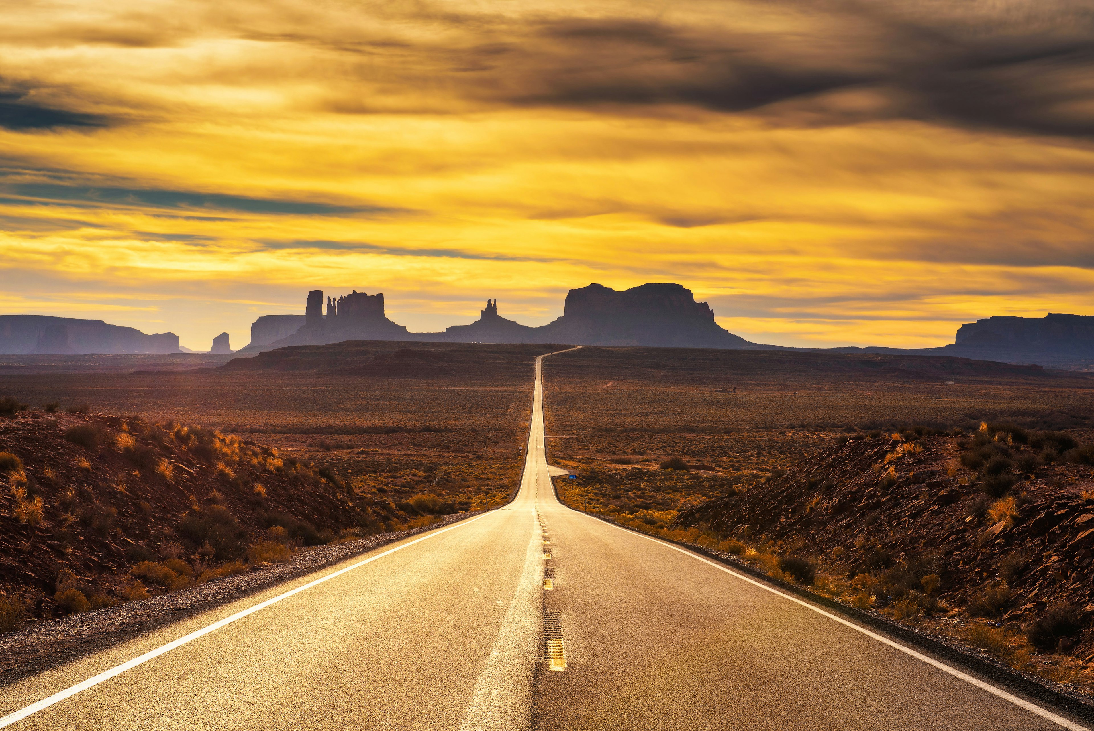 A straight strech of an empty highway leading through the desert at dusk, with the mesas and rock formations silhouetted in the distance, Arizona