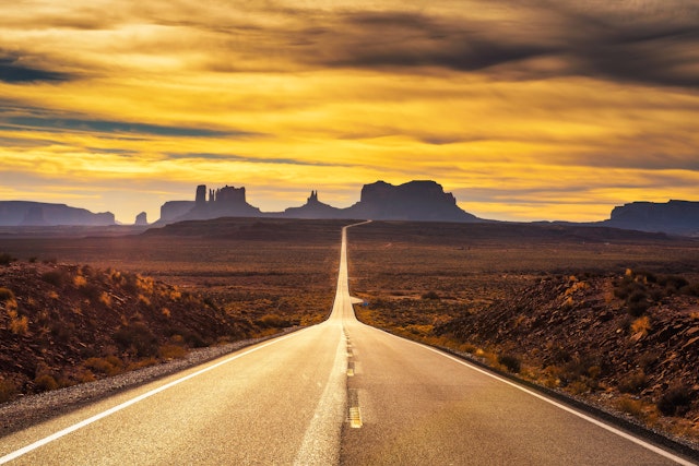 A straight strech of an empty highway leading through the desert at dusk, with the mesas and rock formations silhouetted in the distance, Arizona