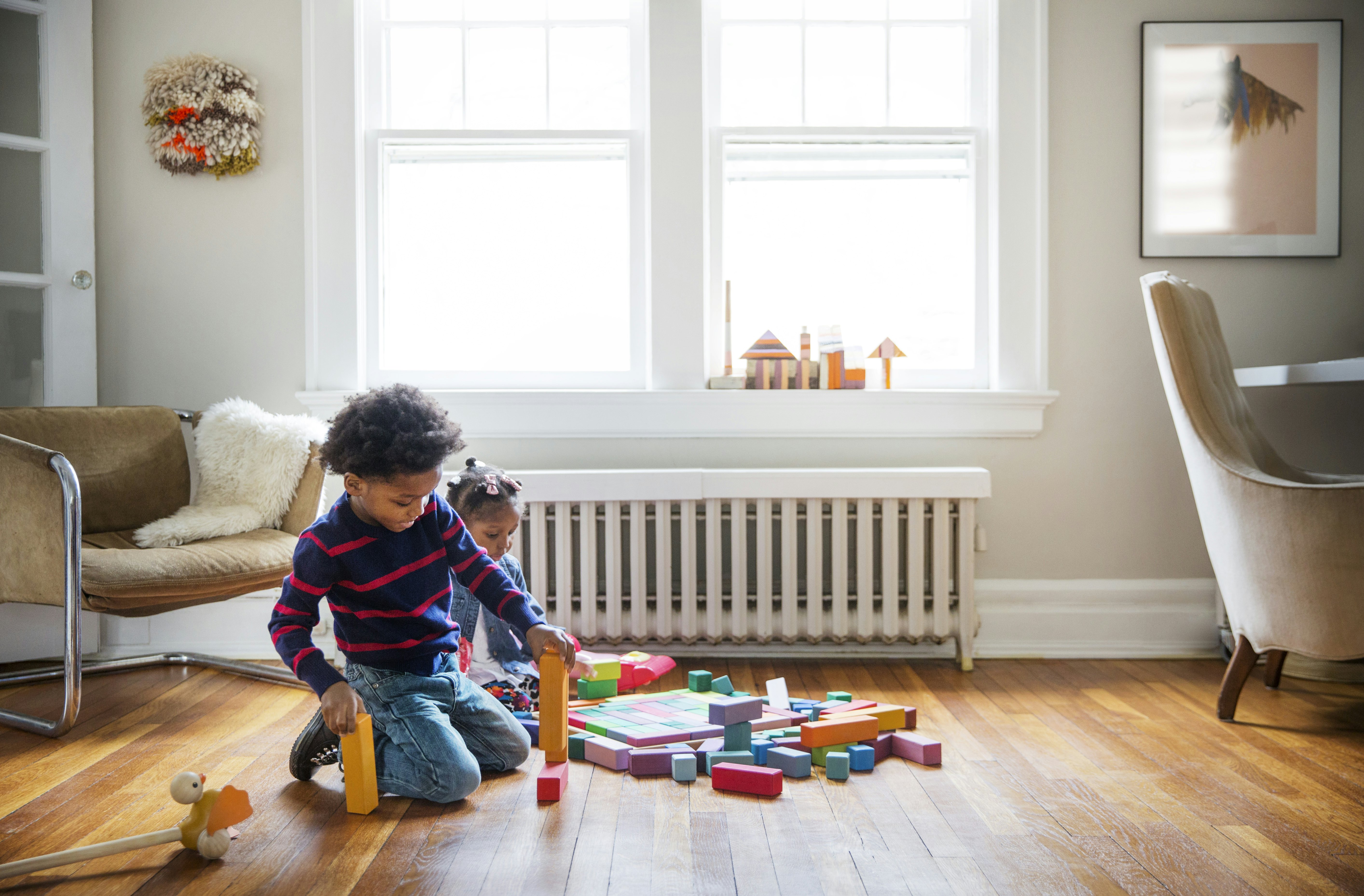 Two young children sit on a wooden floor surrounded by colourful blocks. The oldest child has built a tower