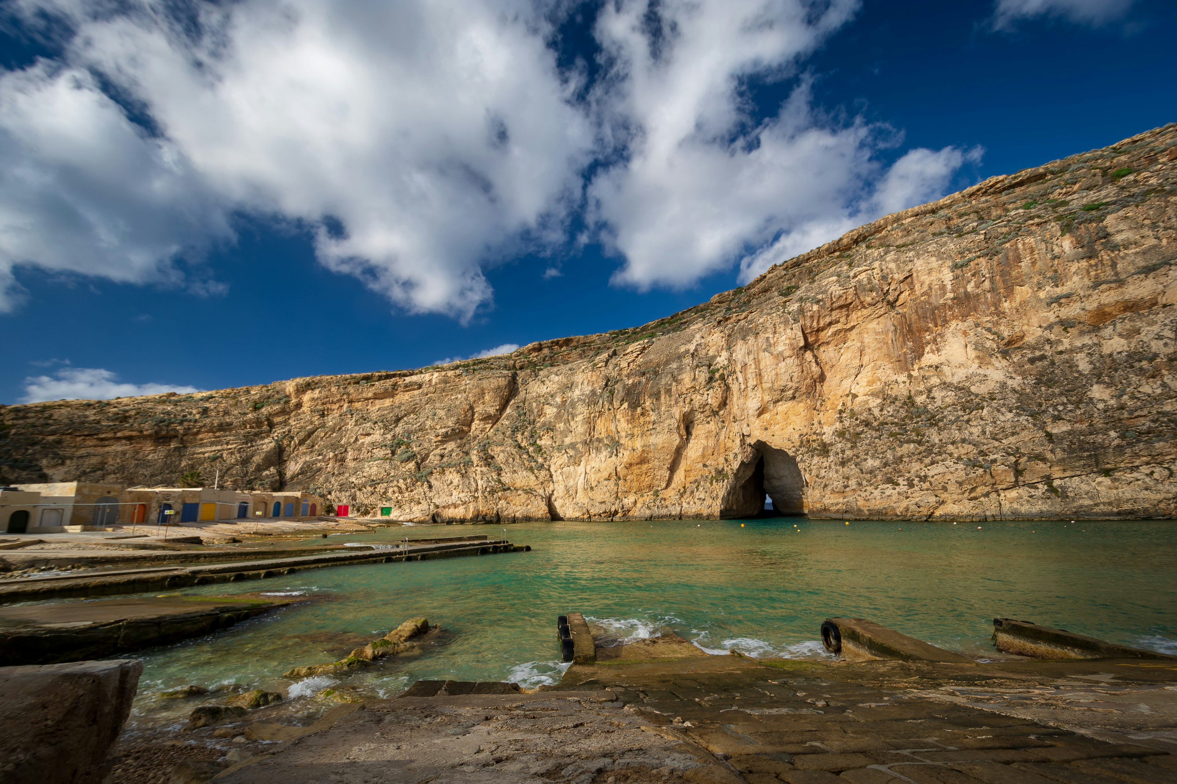 Looking across the turquoise waters of the small Inland Sea the cave entrance to the Med is visible at the base of the rock cliff. Colouful boat houses sit on the shore to the left