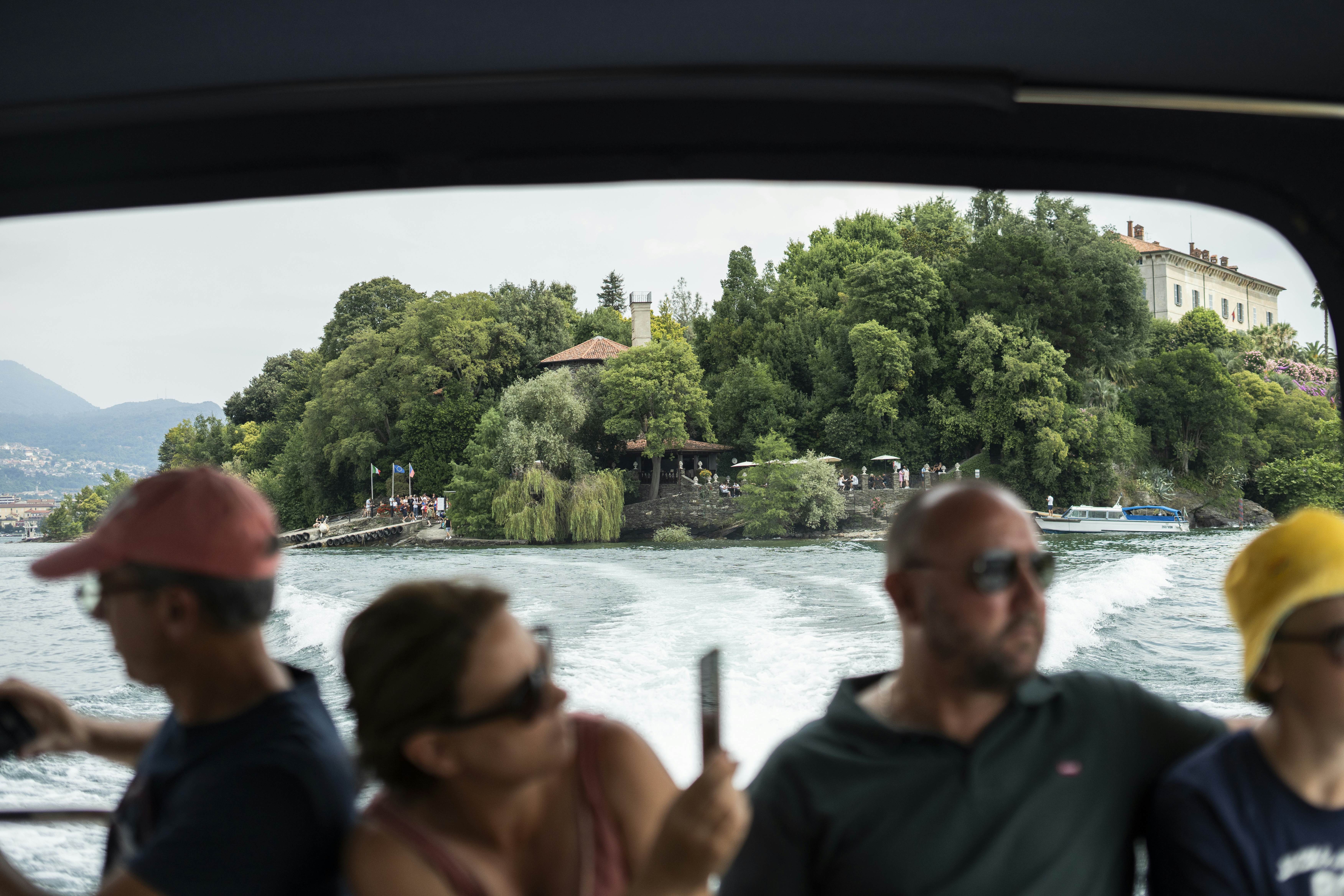 People take photos from a boat as they cruise away from a tree- and villa-covered island.