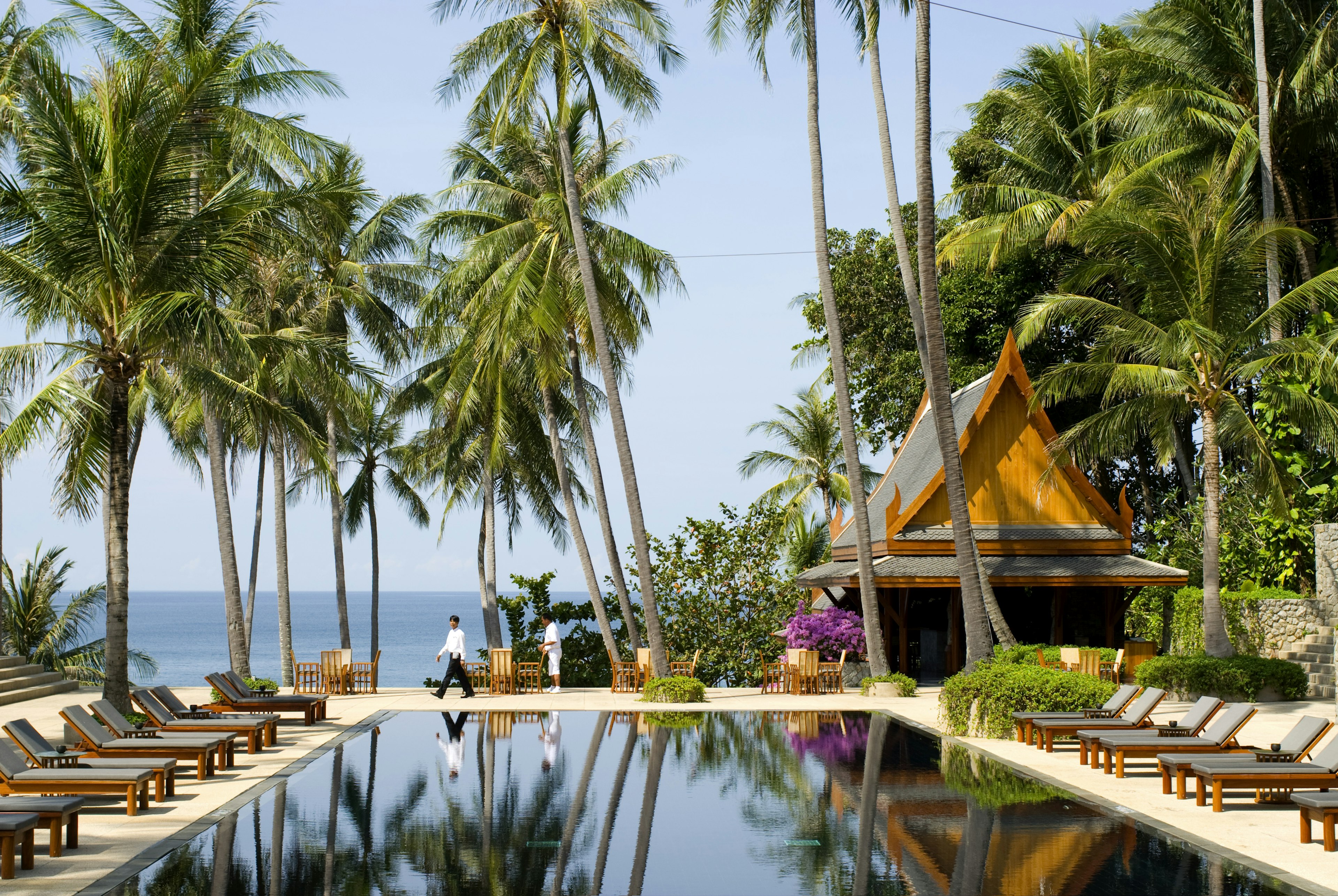 An empty swimming pool, surrounded by wooden deck chairs and palm trees. The sea is in the background as are two staff members.