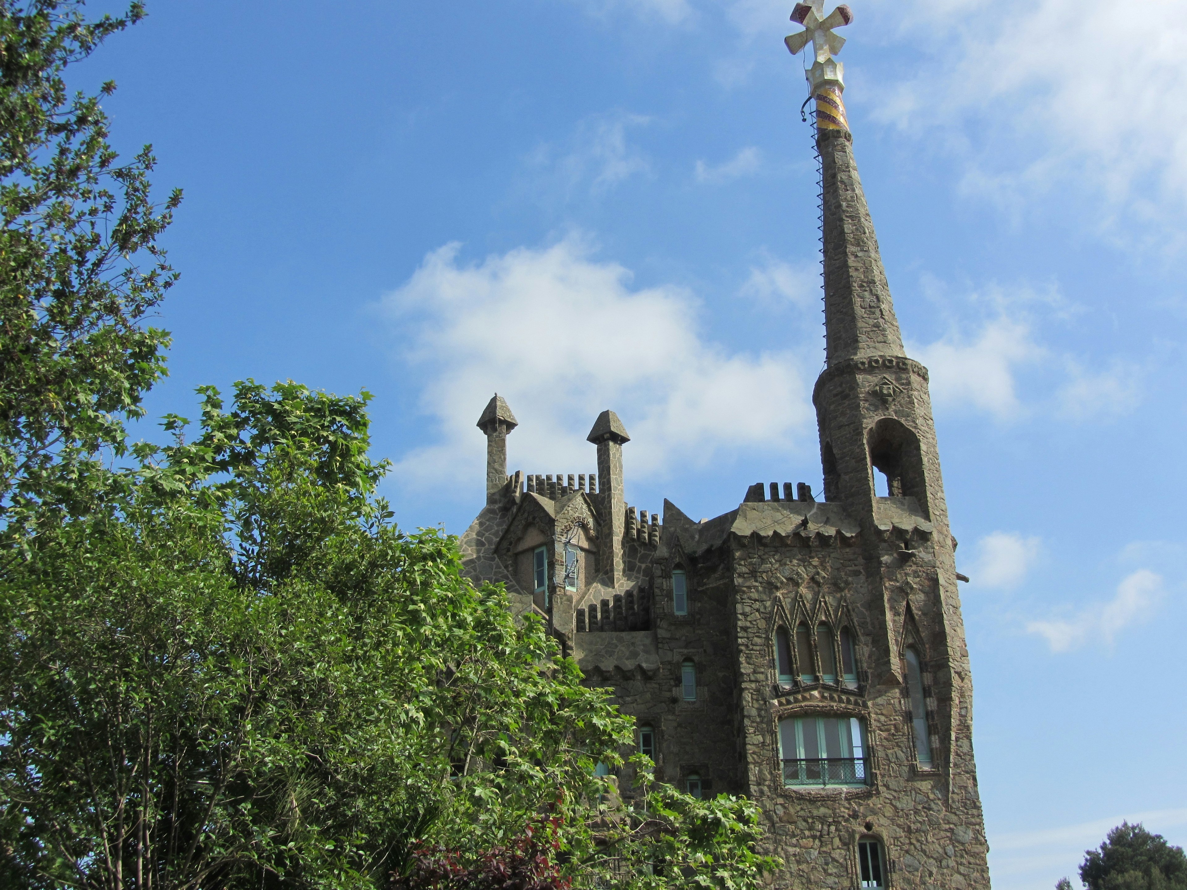 Bellesguard: it has a castle-like appearance with crenellated walls of stone and brick, narrow stained-glass windows, elaborate ironwork and a soaring turret mounted by a Gaudían cross. There are trees in its grounds.