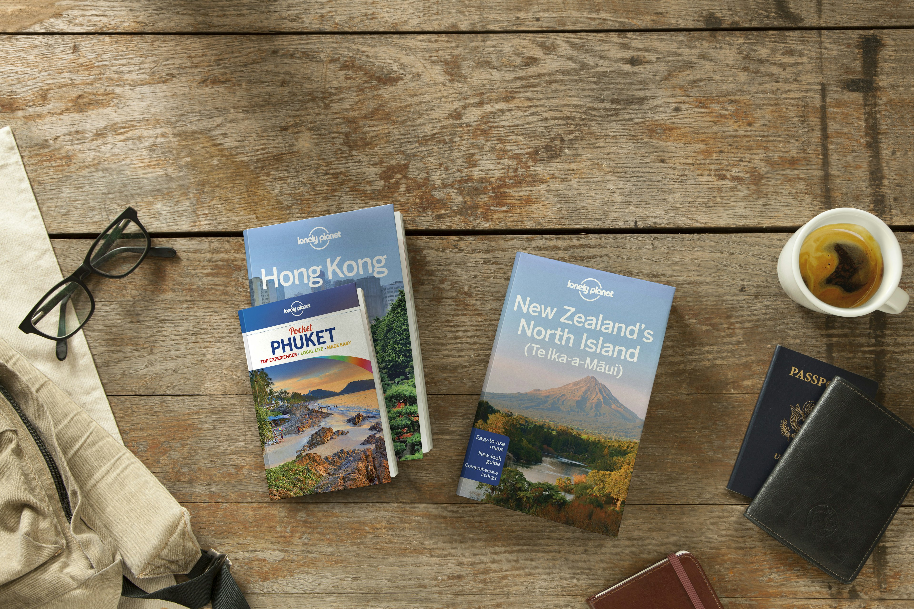 Several Lonely Planet guidebooks sit on a warm, honey-hued wooden table with large planks. One is a guide to Hong Kong, another to Phuket. A pair of black eye glasses sit off to the side. Another is a guide to the north islands of New Zealand, sitting by a blue American passport and a mug of herbal tea.