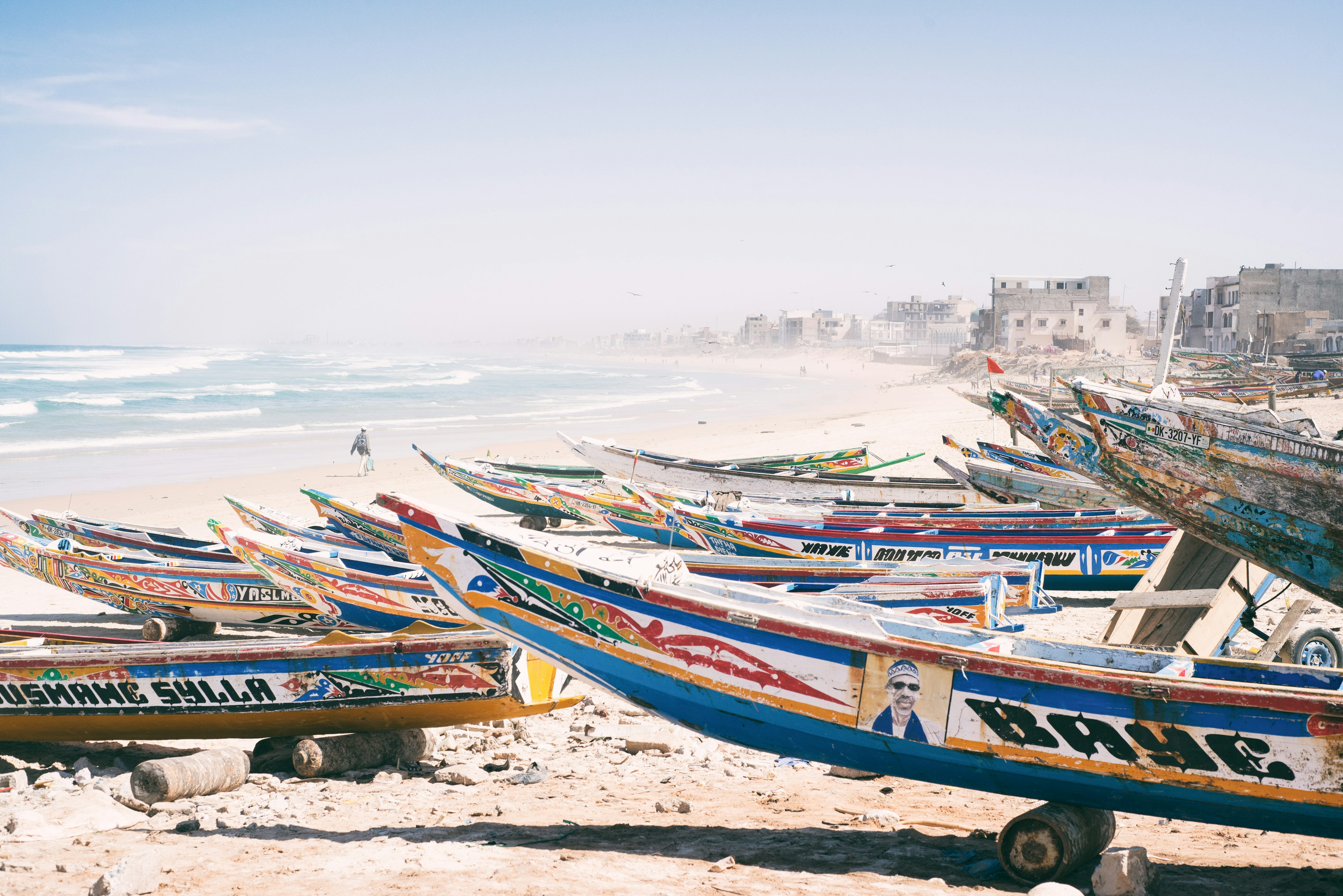 Long canoes painted in vibrant blues, yellows and reds sit side-by-side on the beach; waves roll in, while cement buildings back the scene.