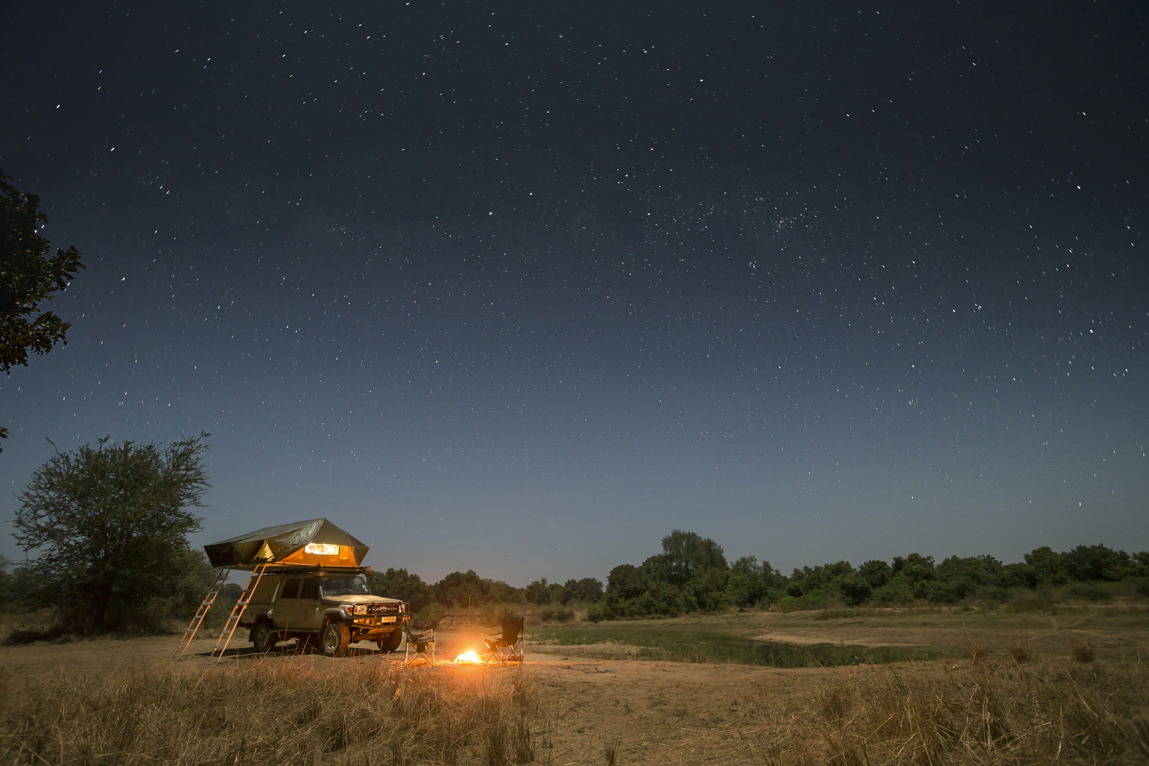 A tent set up at the back of a vehicle with a small campfire burning in front. The night sky is full of stars above it.