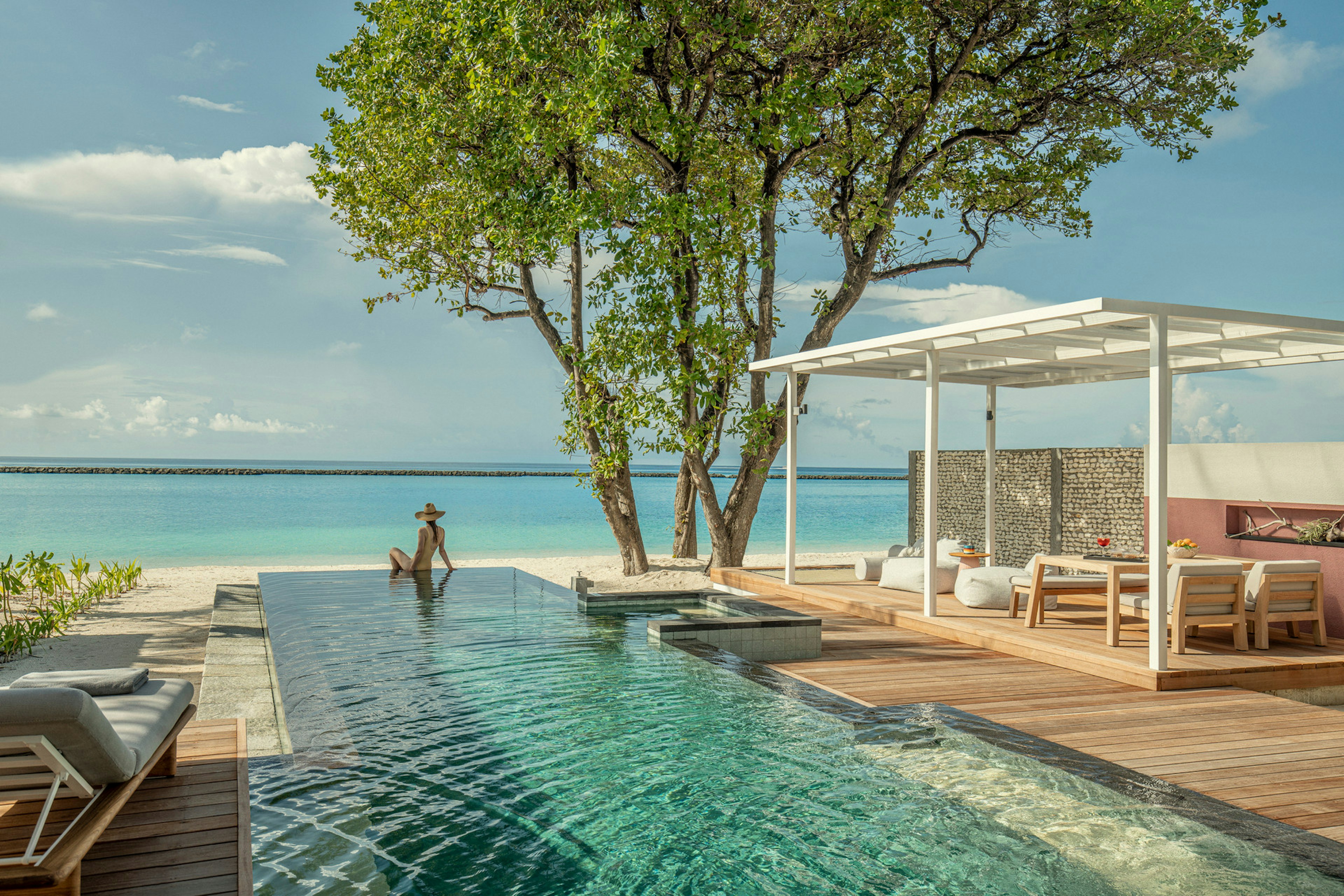 A woman in a sun hat sits at the edge of an infinity pool looking out at the ocean