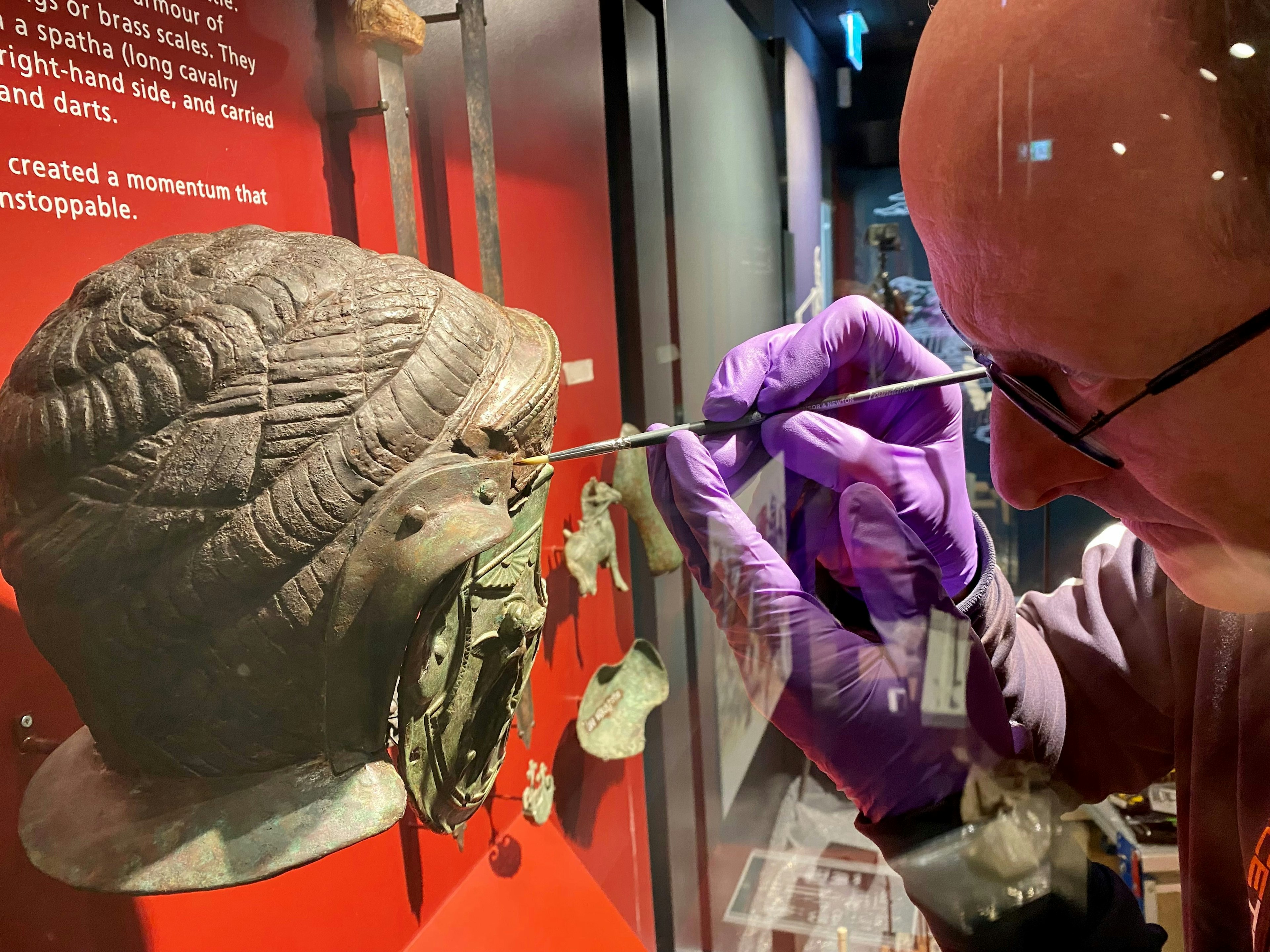 A museum curator cleans a display of Roman artifacts including a helmet