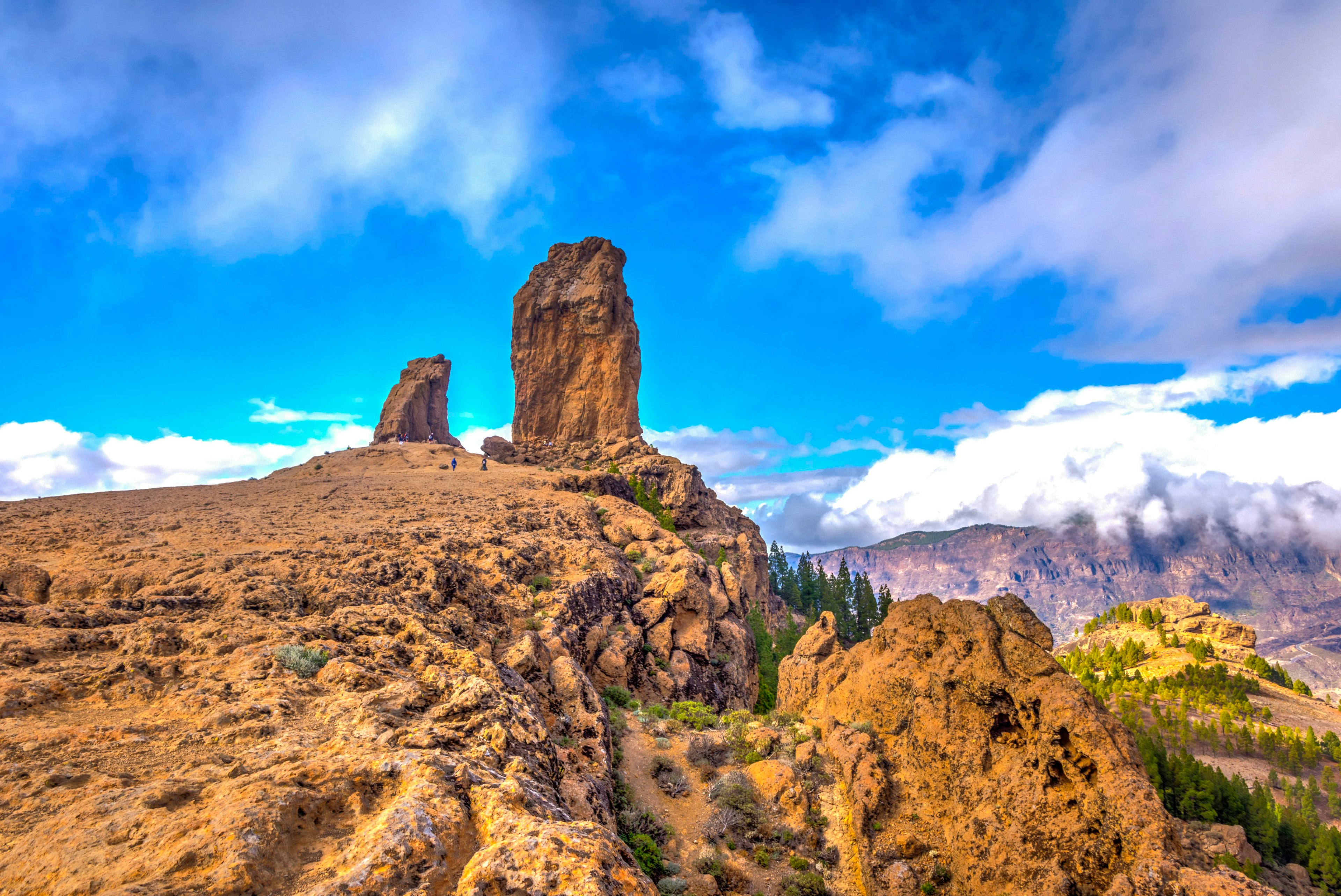 Two tall rock formations stand at the top of a hill with more cloud-covered mountains in the distance