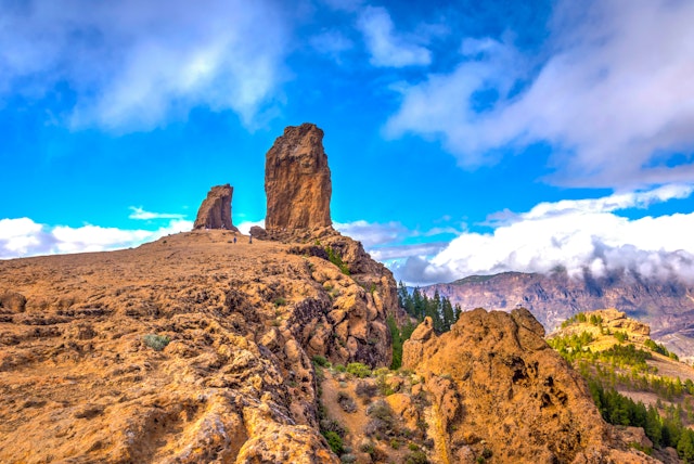 Two tall rock formations stand at the top of a hill with more cloud-covered mountains in the distance