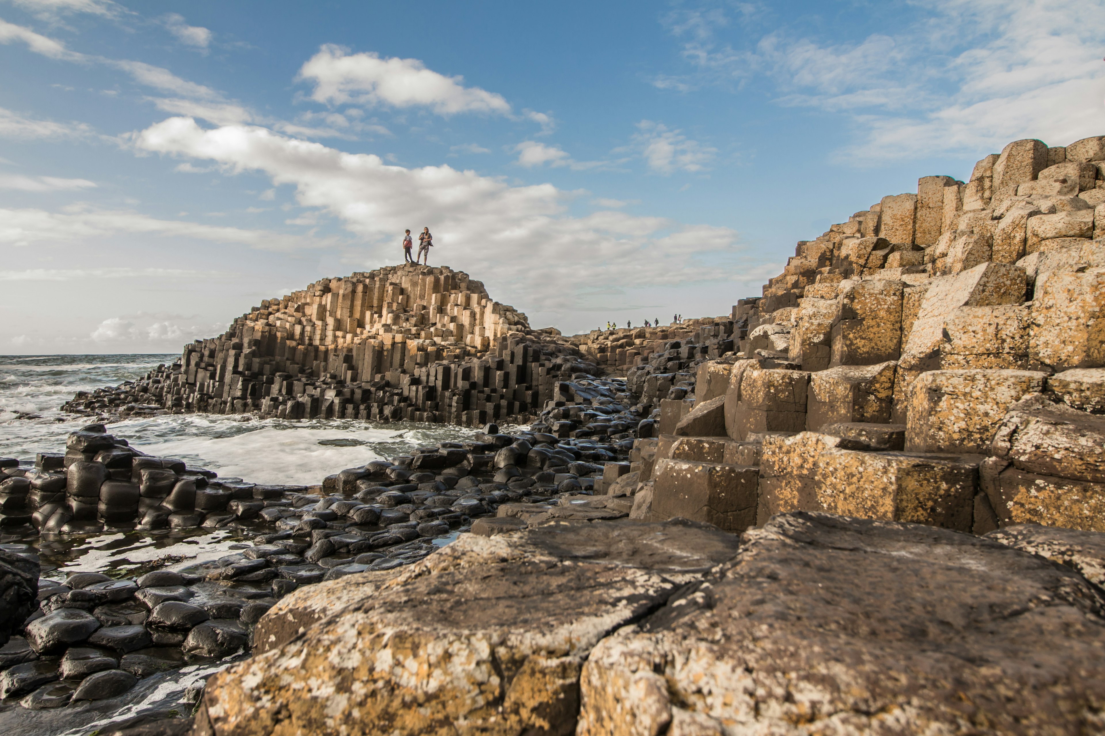 Two people walk along the rising basalt columns of the Giant's Causeway, with the sea in the background.