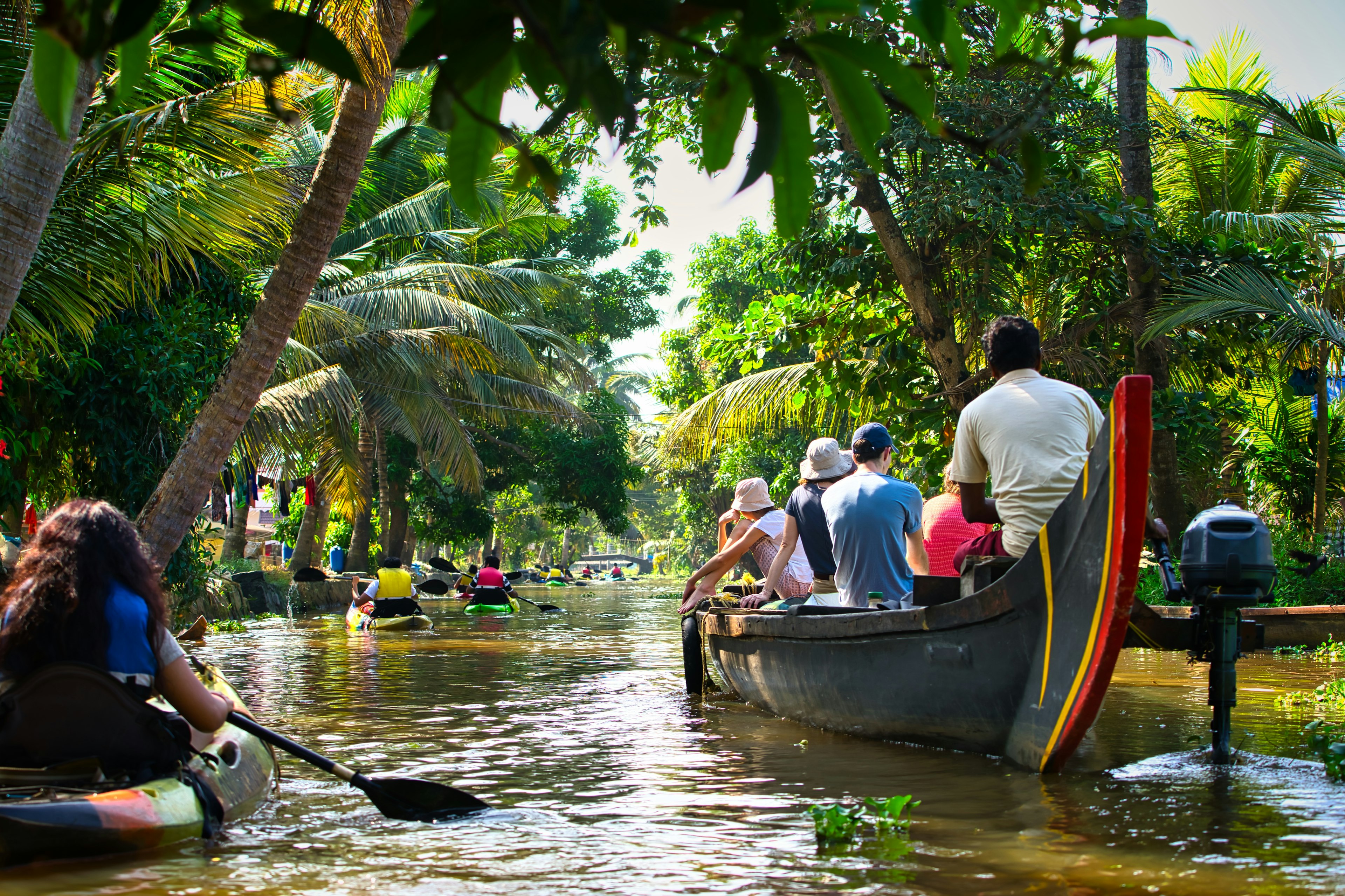 Tourists paddling in boats or kayaks on backwaters of Kerala, surrounded by wilderness and trees, in the bright sunlight, in the middle of a beautiful day.