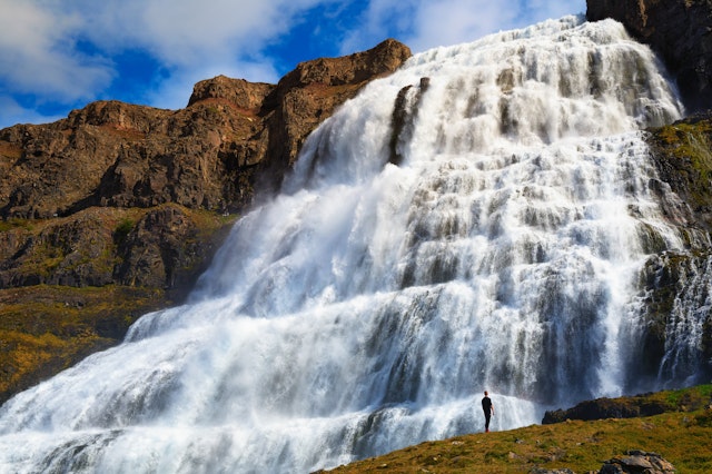 A person stands at the base of a vast waterfall that cascades down a rippled rock face
