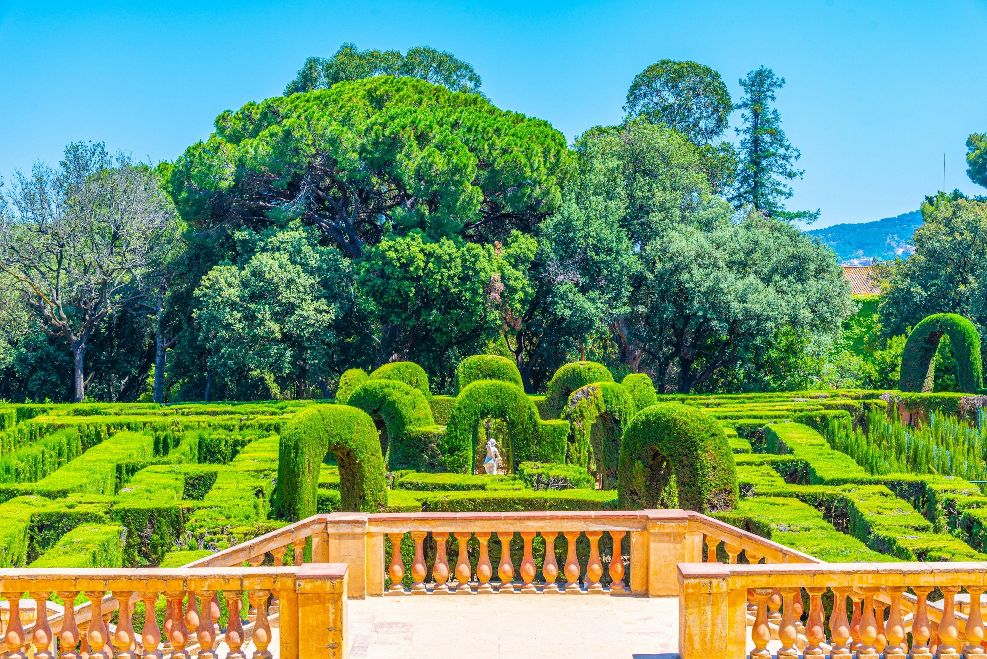 Looking over a pillared wall to a cypress hedge labyrinth below, with trees beyond.