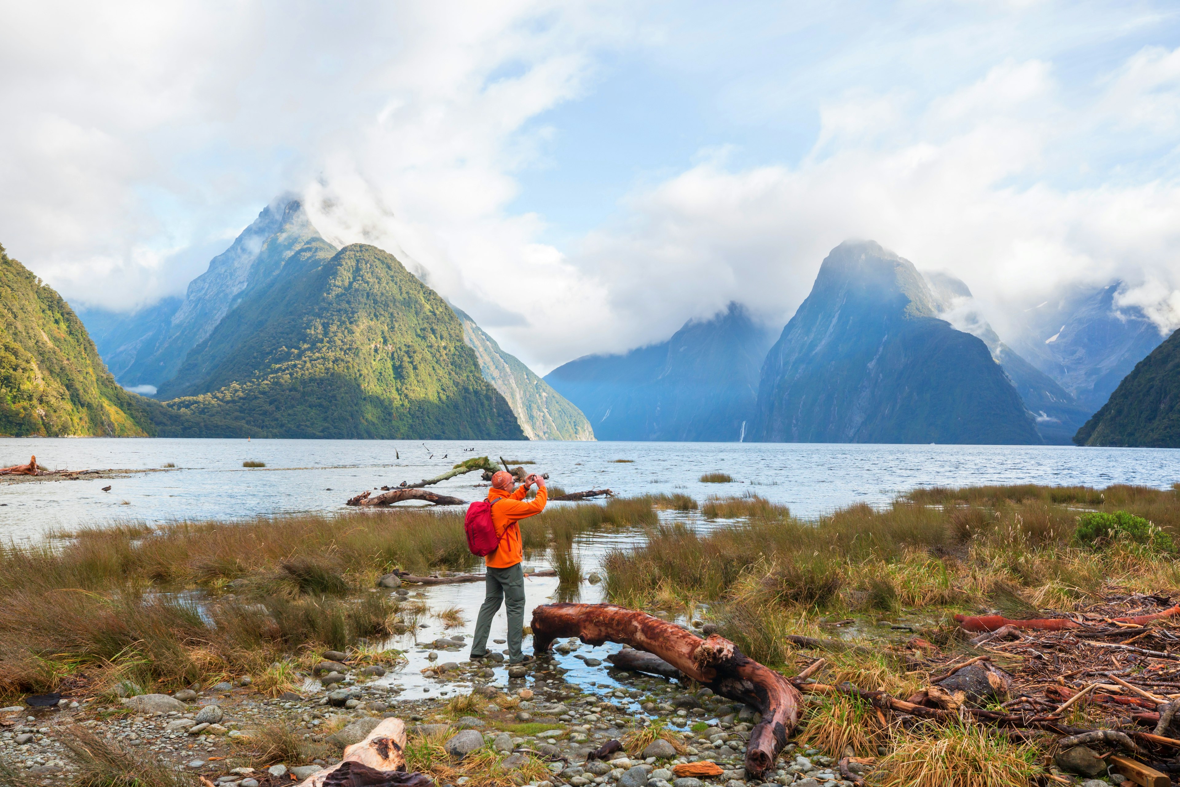 A tourist in red takes a photo of the beautiful hills and water of Milford Sound in New Zealand.