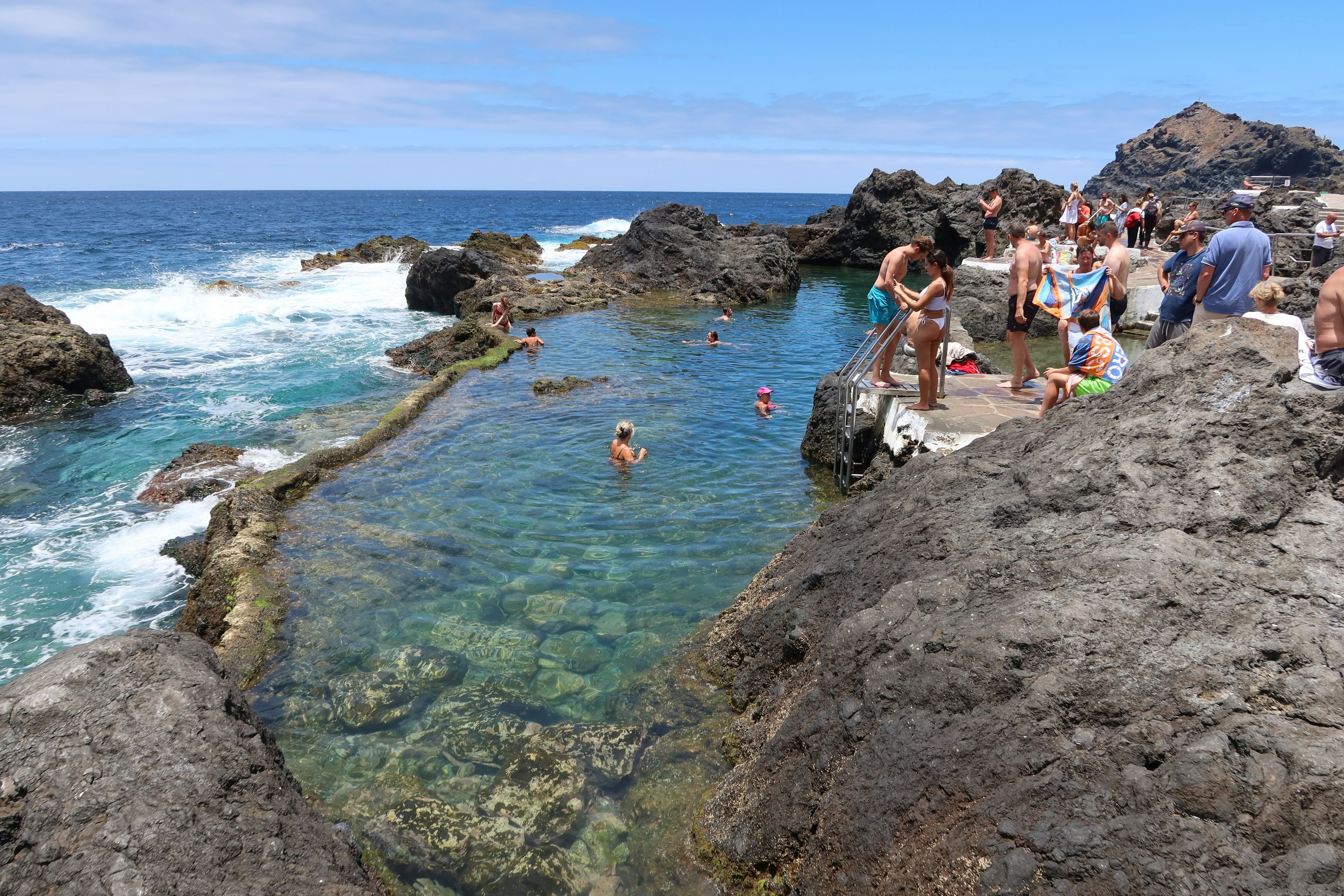 People swim in tidal pools next to the crashing surf on a shoreline with volcanic rocks