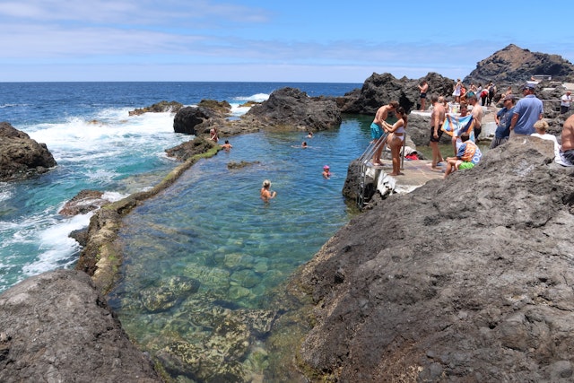 People swim in tidal pools next to the crashing surf on a shoreline with volcanic rocks