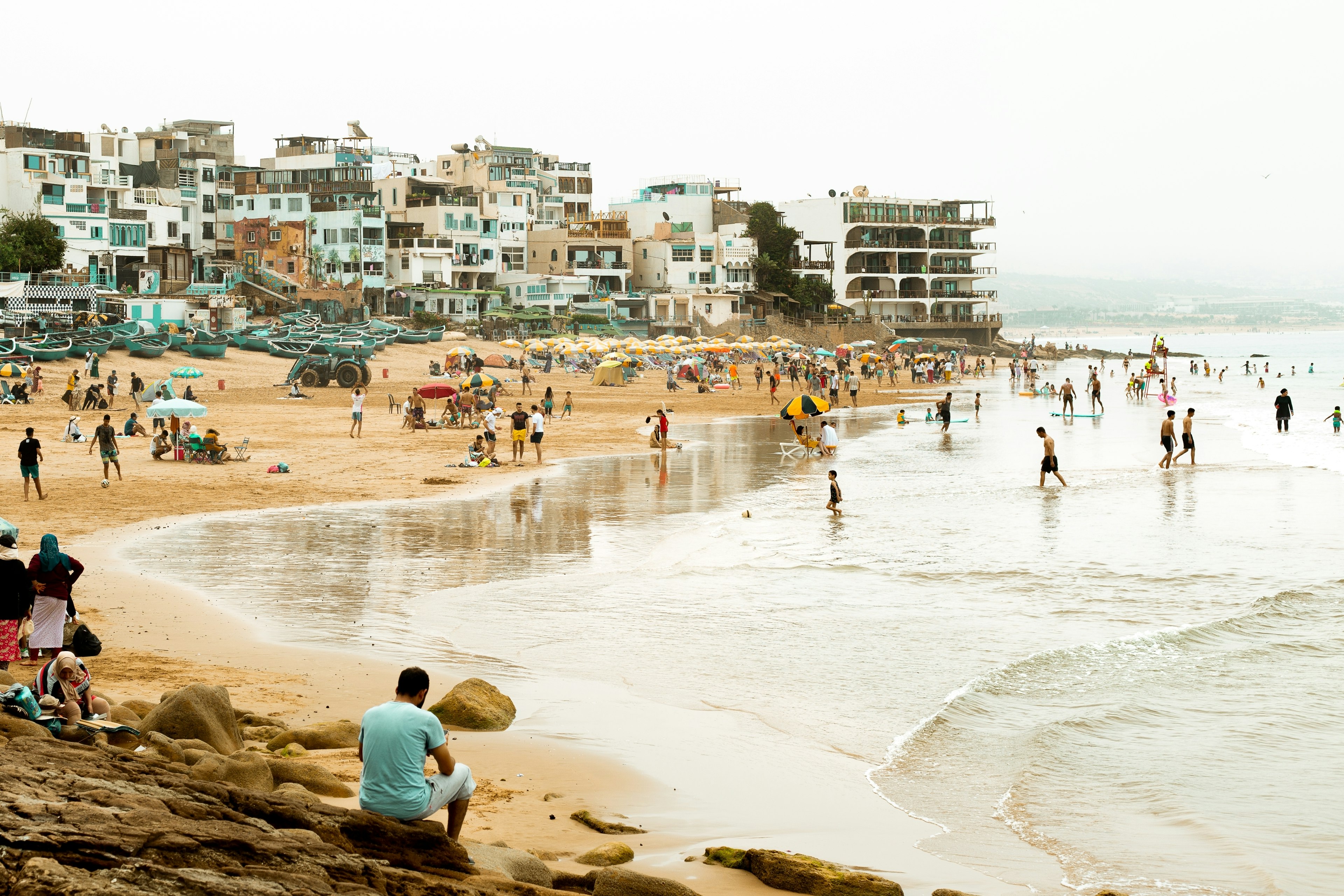 A busy beach on a grey day with people walking in the water and apartments in the background.