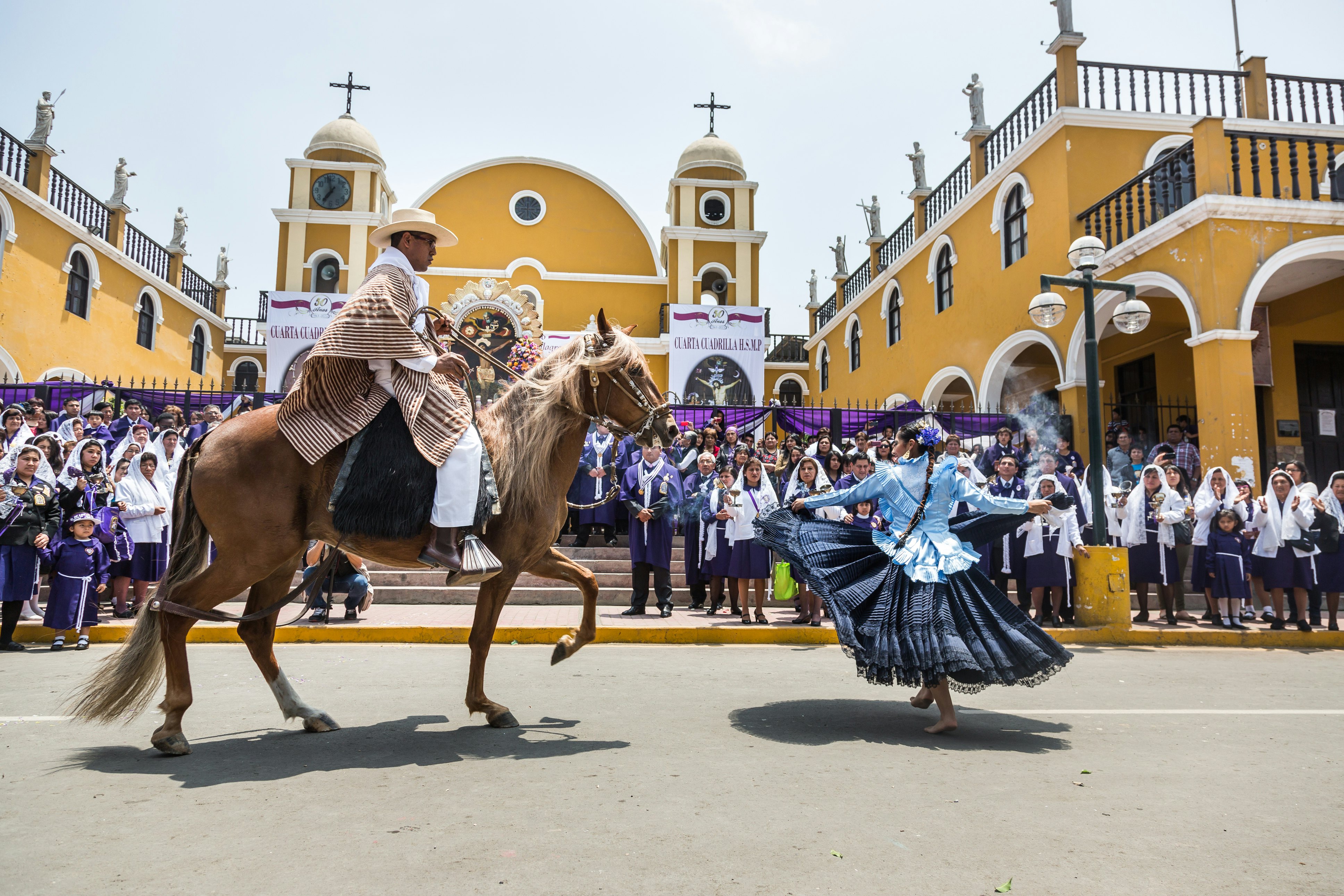 A man wearing a striped poncho rides a horse behind a woman wearing a full blue skirt; they are in front of a crowd of people standing by a yellow building with crosses on two towers.