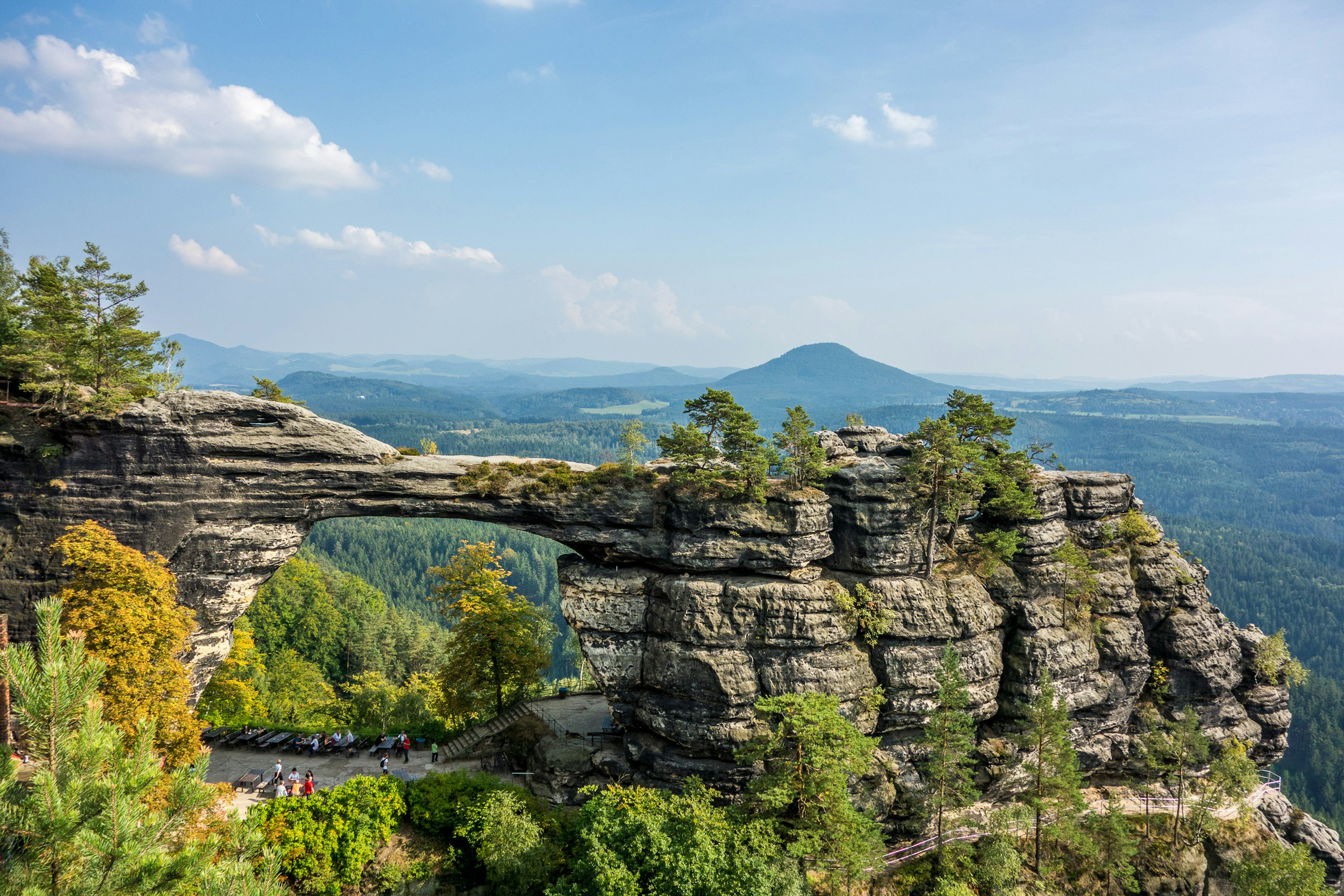 A natural sandstone arch rock formation surrounded by trees, with a view of a wooded expanse and hills in the distance