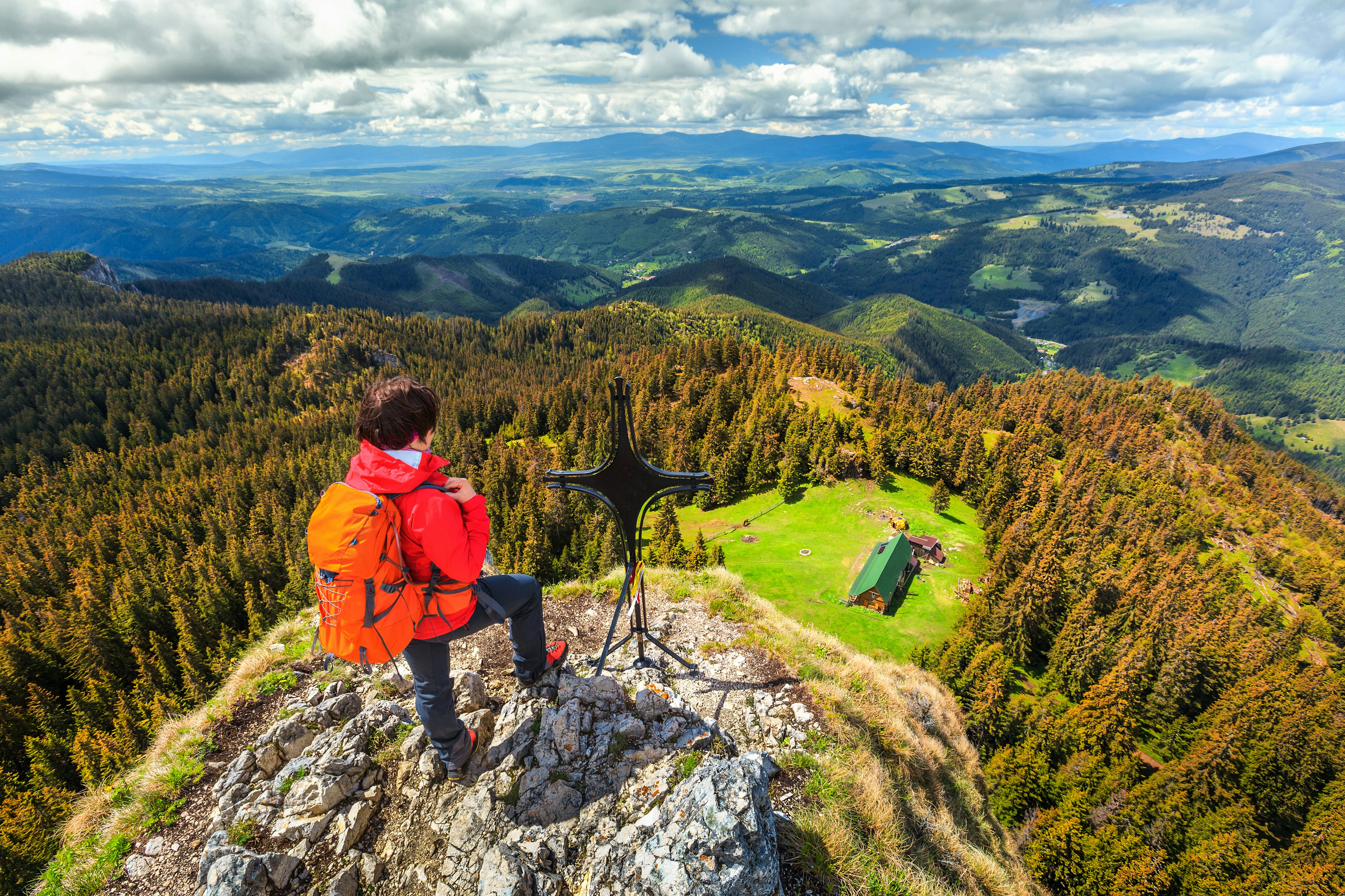 A female hiker with a red parka and orange backpack stands at a rocky outcrop with a cross, looking out at hills with trees showing autumn foliage