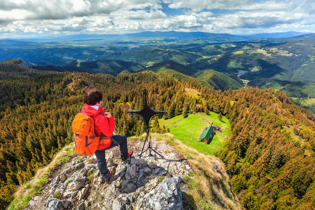 A female hiker with a red parka and orange backpack stands at a rocky outcrop with a cross, looking out at hills with trees showing autumn foliage