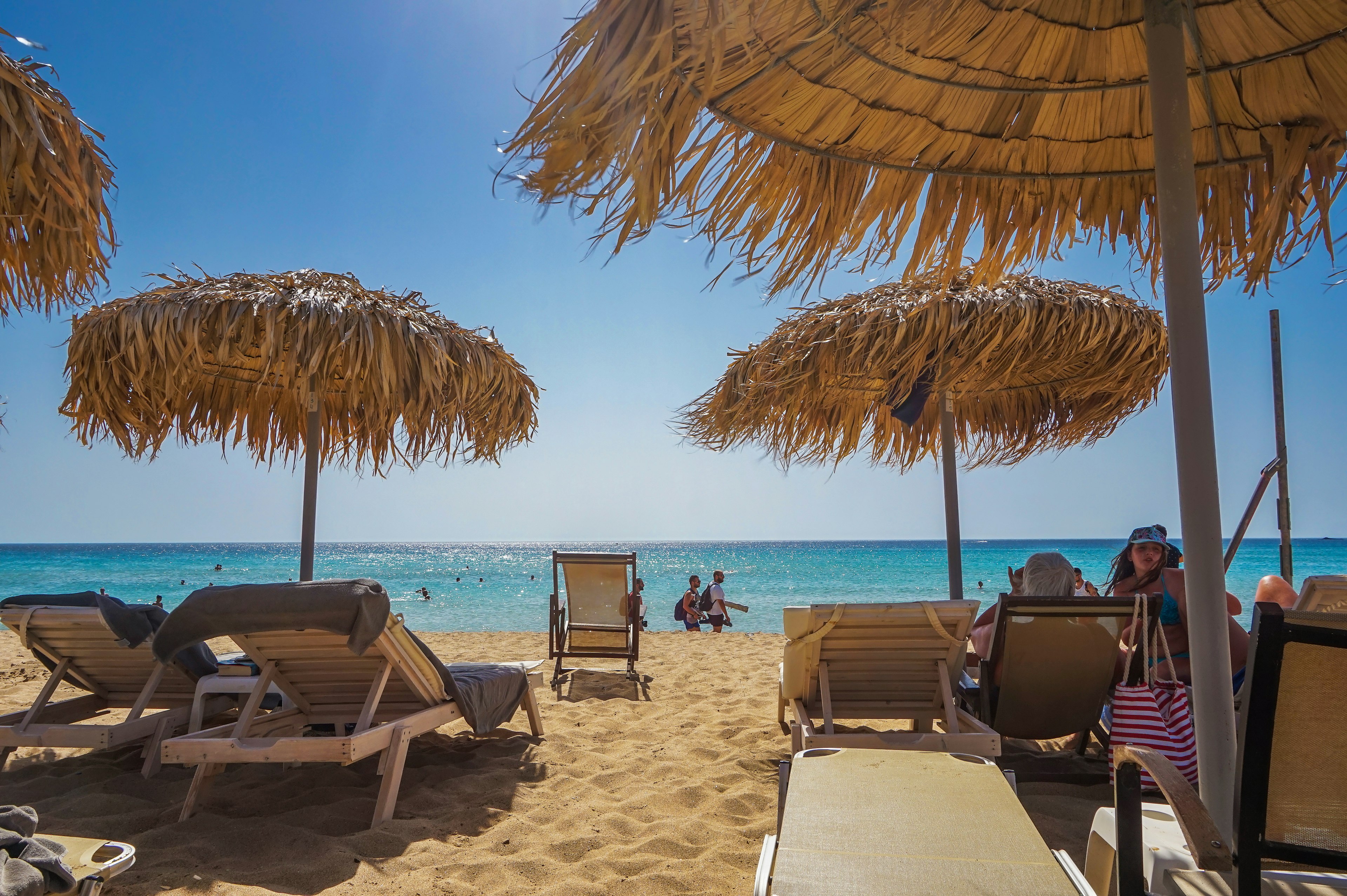 A sandy beach in Crete with sunbathers in chaise lounge and swimmers in the ocean.