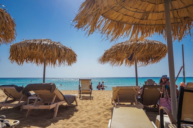 A sandy beach in Crete with sunbathers in chaise lounge and swimmers in the ocean.