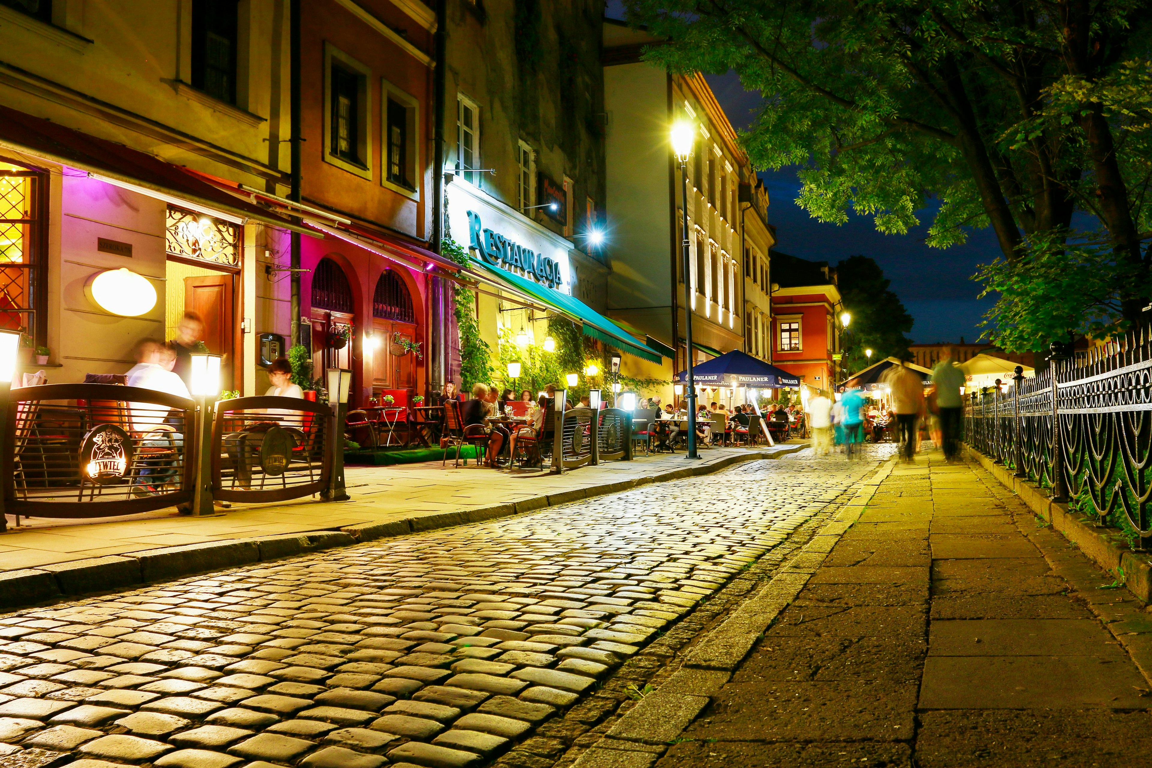 A view of cobblestones on a narrow street with the brightly illuminated facades of restaurants at night