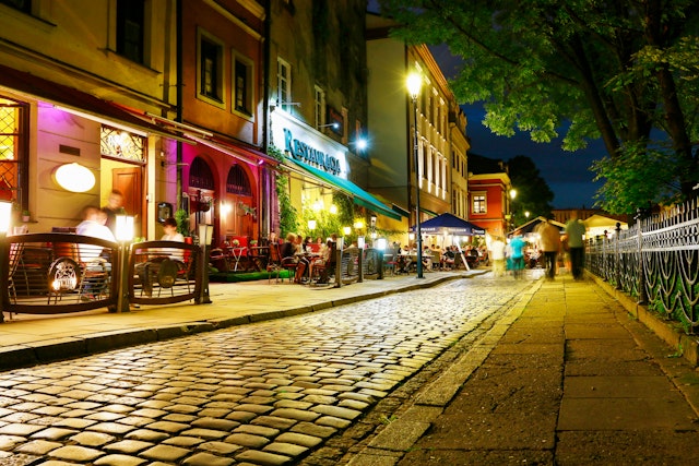 A view of cobblestones on a narrow street with the brightly illuminated facades of restaurants at night