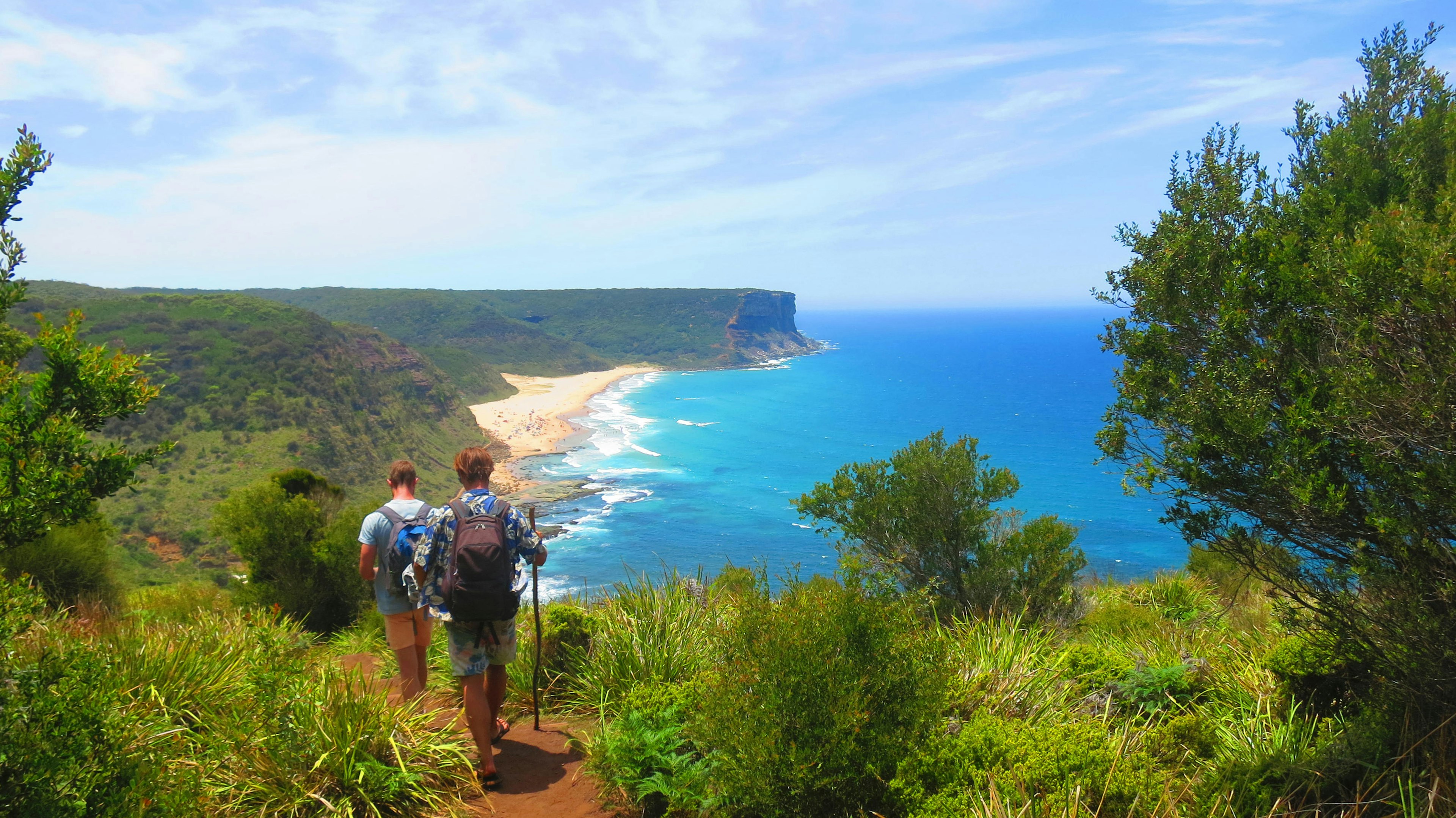 Two young men with with sticks and backpacks hike on a narrow path through lush bushes with a view of the ocean, cliffs and beaches in the distance