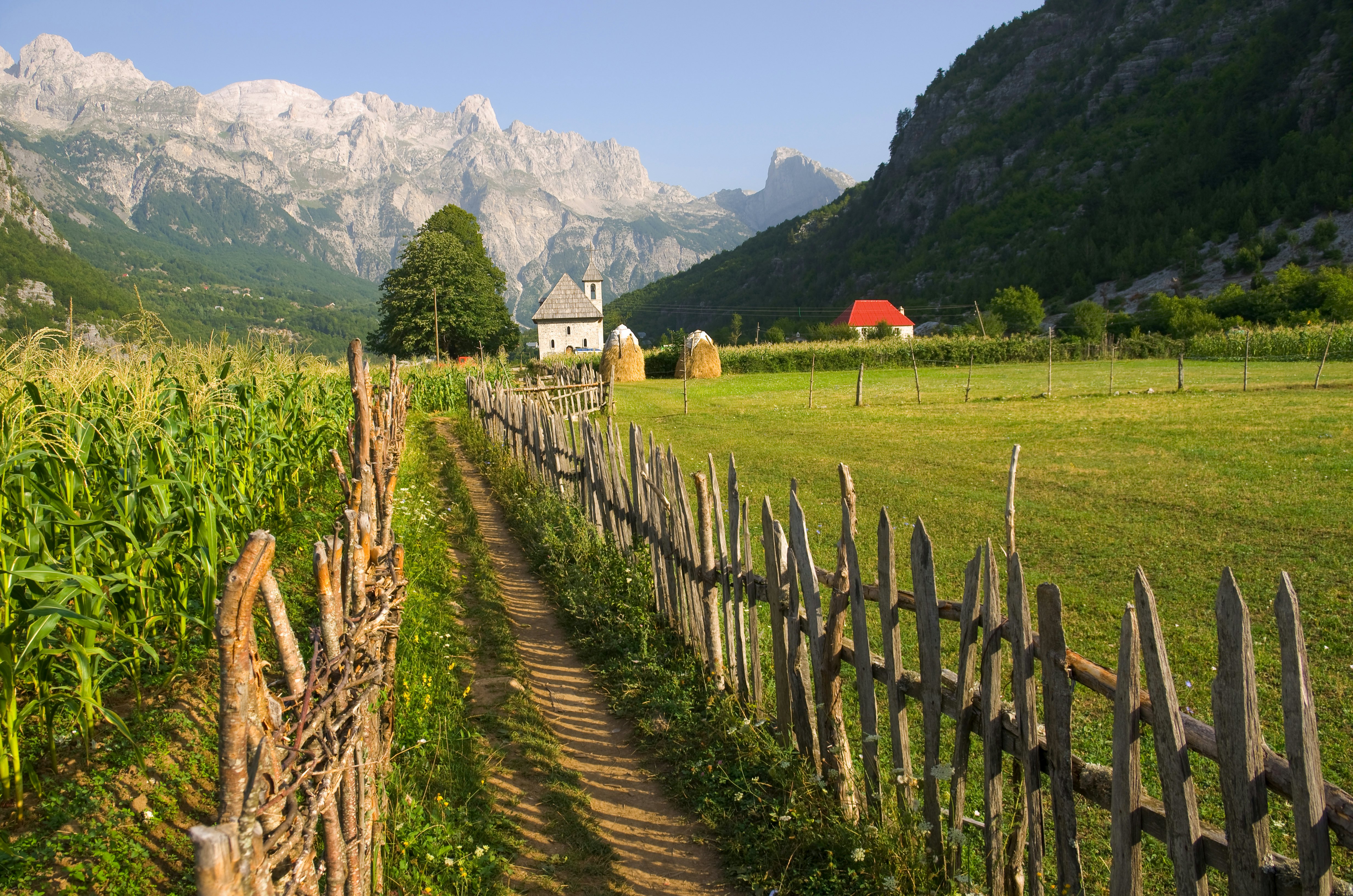 a path with wooden fence leads the catholic church in the Theth Valley, on background the mountains of Albanian Alps